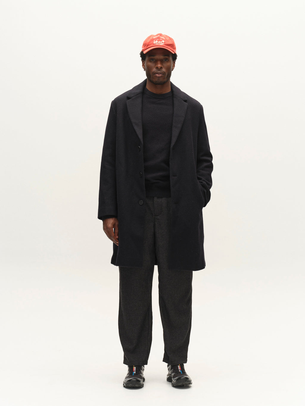 A man stands against a plain background wearing an effortlessly stylish look: the Gabba GABleeds Plain Coat in navy layered over a dark sweater, paired with grey pants, black shoes, and a red cap. One hand rests in his pocket as he gazes straight ahead, perfectly capturing modern everyday wear.