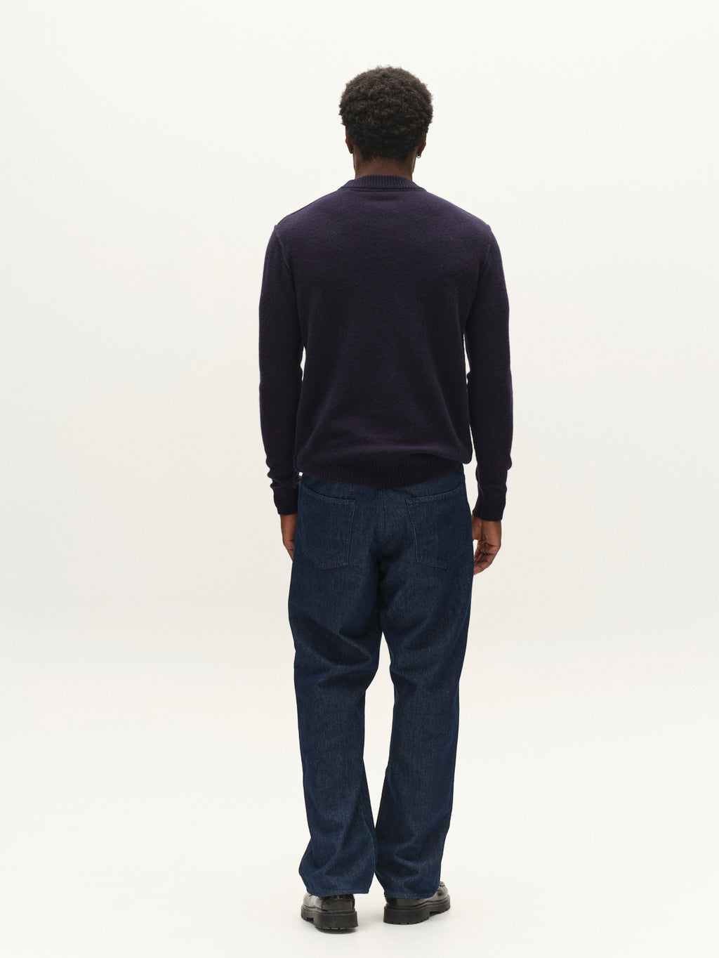 A person stands facing away from the camera, dressed in the Gabba GABtokota Element Aico LS Knit sweater in navy and blue jeans against a plain, light background, embodying everyday comfort.