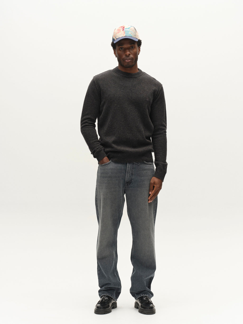 A man wearing the Gabba GABtokota Element Aico LS Knit crew neck sweater in Dark Grey Mel, paired with faded jeans, black shoes, and a colorful cap stands against a plain white background with one hand in his pocket.