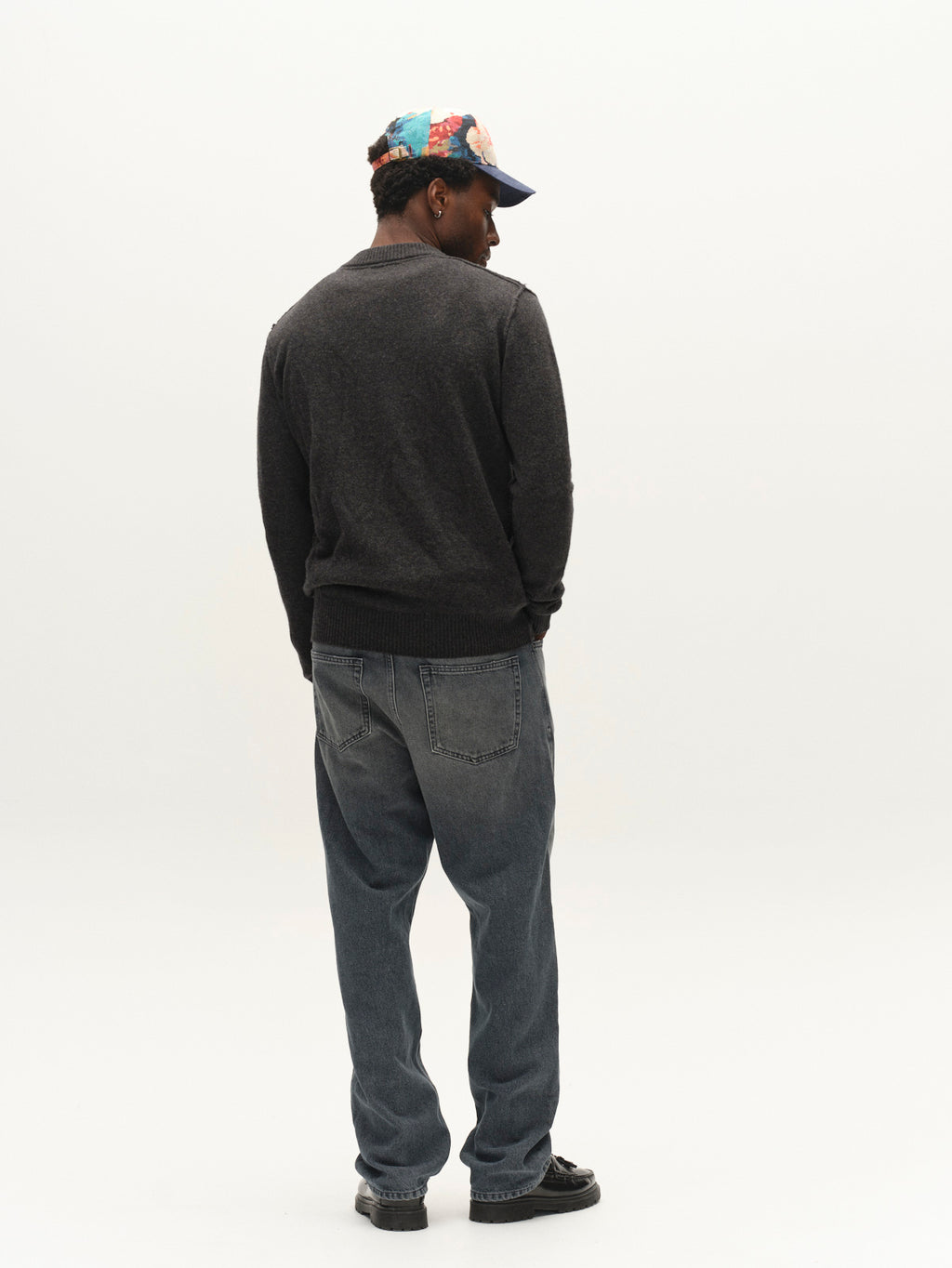A person stands facing away, wearing a Gabba GABtokota Element Aico LS Knit in Dark Grey Mel., blue jeans, black shoes, and a blue cap with a floral pattern, against a plain white background.