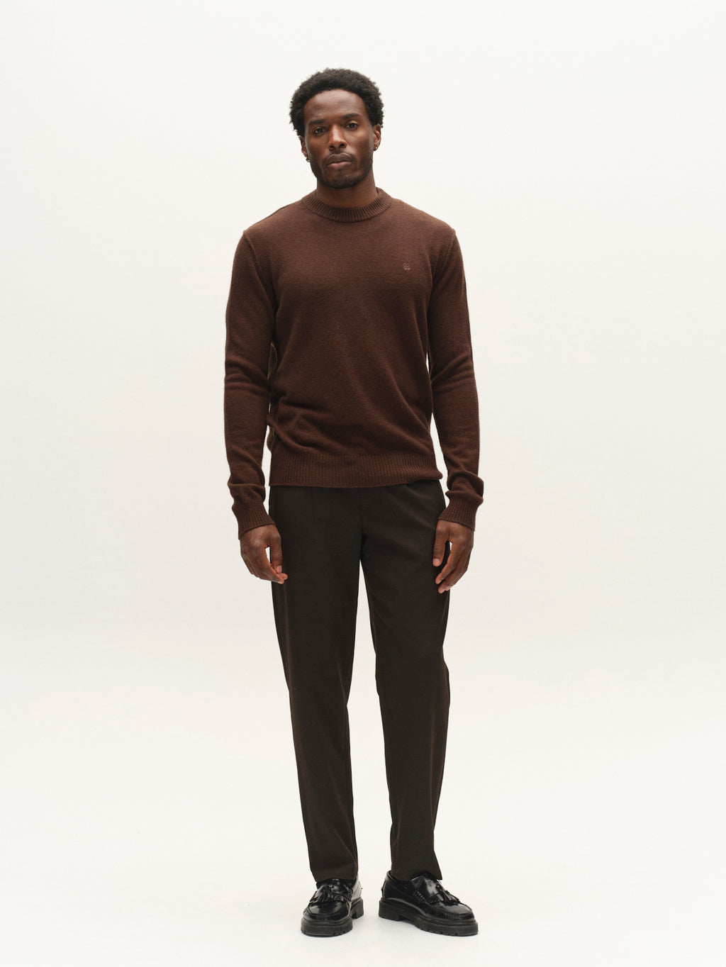 A man stands against a plain white background, wearing the Gabba GABtokota Element Aico LS Knit in Brown Slate, paired with dark brown pants and black shoes. He gazes forward with a neutral expression, his short hair complementing the timeless comfort and style of his classic Gabba outfit.
