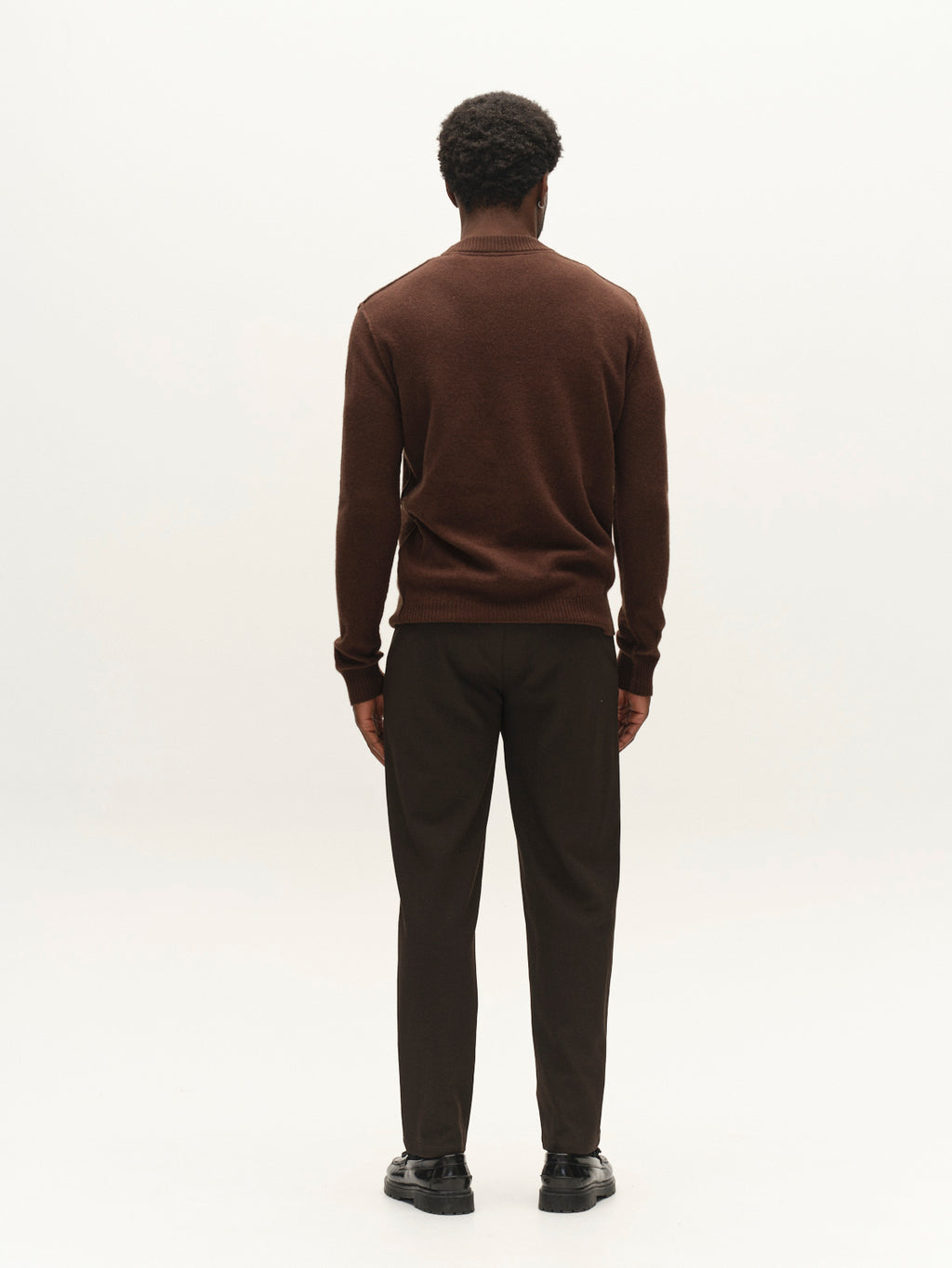 A man stands with his back to the camera, wearing the Gabba GABtokota Element Aico LS Knit in Brown Slate—a soft wool crew neck sweater designed for everyday comfort—paired with dark brown pants and black shoes against a plain white background.