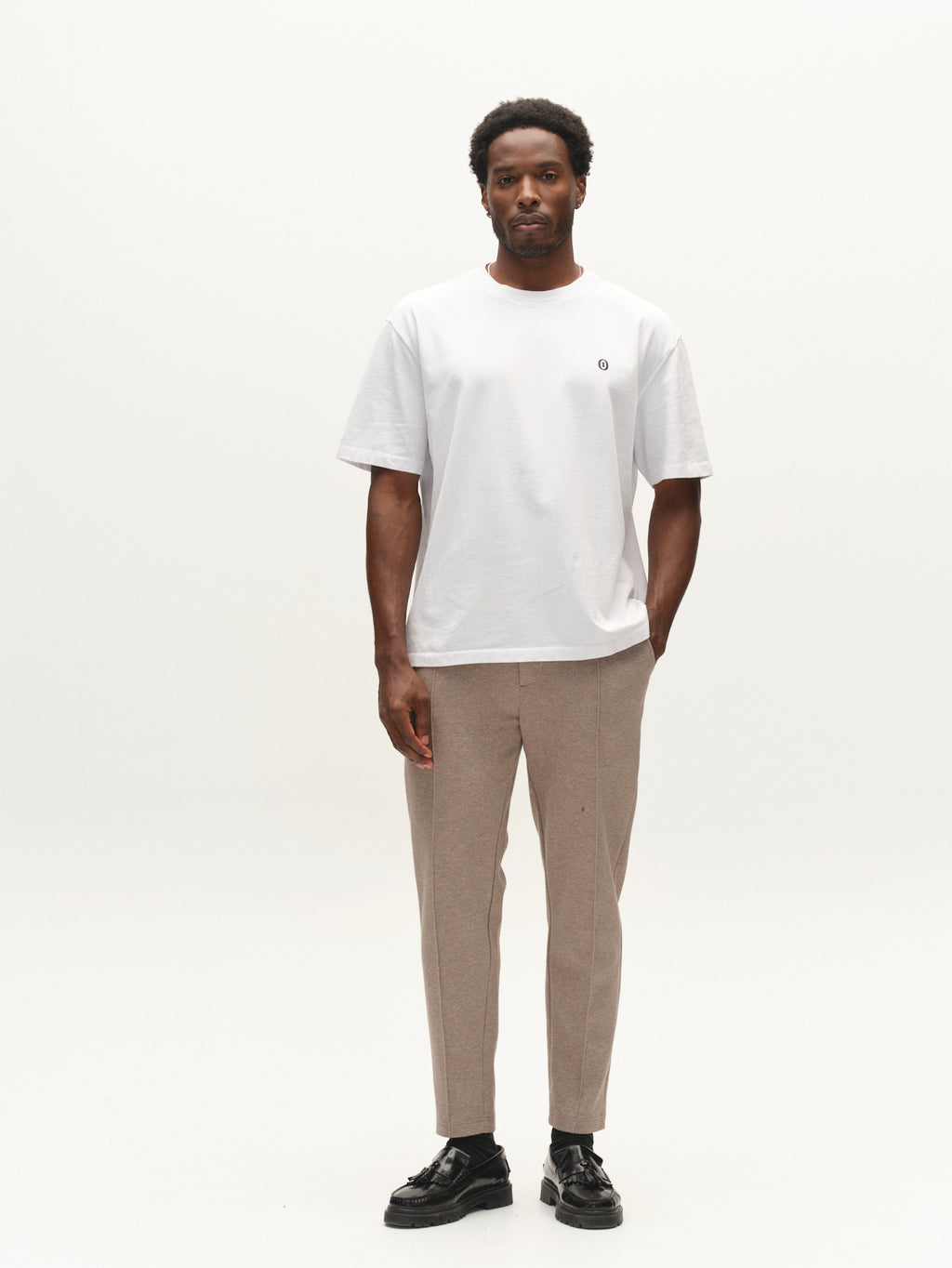 A man stands against a plain white background, wearing the Gabba GABlegin Wix Boxy Hour Lobby1 SS T-shirt in white, paired with beige pants and black shoes. His arms are relaxed by his sides as he looks directly at the camera.
