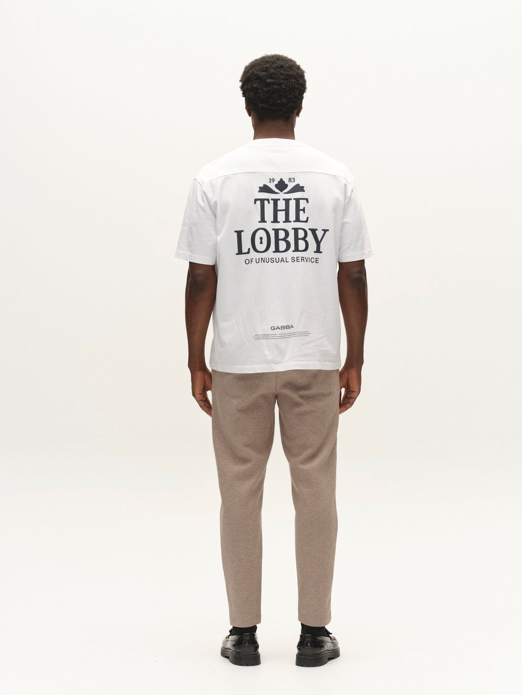 A person stands facing away, wearing the Gabba GABlegin Wix Boxy Hour Lobby1 SS T-shirt in white—a relaxed, boxy-fit tee crafted from textured cotton jersey and featuring THE LOBBY OF UNUSUAL SERVICE as a graphic back print. They pair it with light brown pants and black shoes against a plain white background.