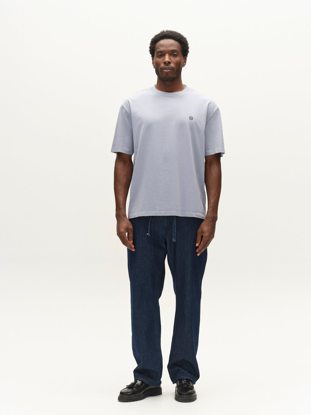 A man stands facing forward, wearing the Gabba GABlegin Wix Boxy Hour Lobby1 SS T-shirt in Dusty Blue, featuring a relaxed boxy fit crafted from textured cotton jersey. He pairs it with dark blue pants and black shoes, set against a plain white background.