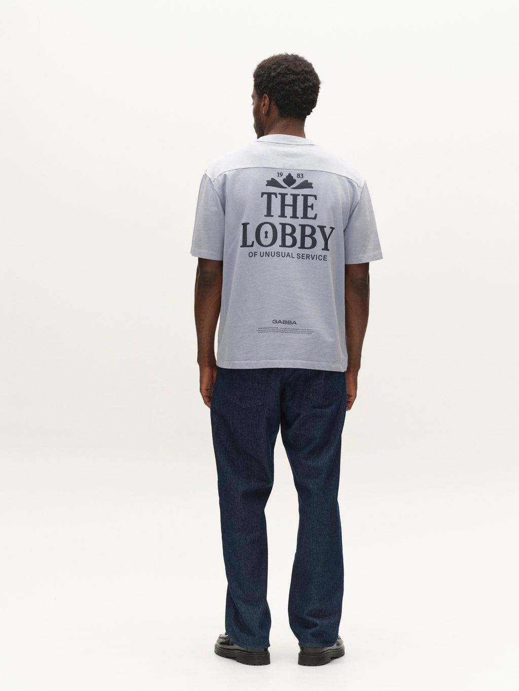 A man stands facing away, wearing the Gabba GABlegin Wix Boxy Hour Lobby1 SS T-shirt in Dusty Blue—a textured cotton jersey top with a relaxed, boxy fit and THE LOBBY OF UNUSUAL SERVICE graphic back print—paired with blue jeans and black shoes against a plain white background.