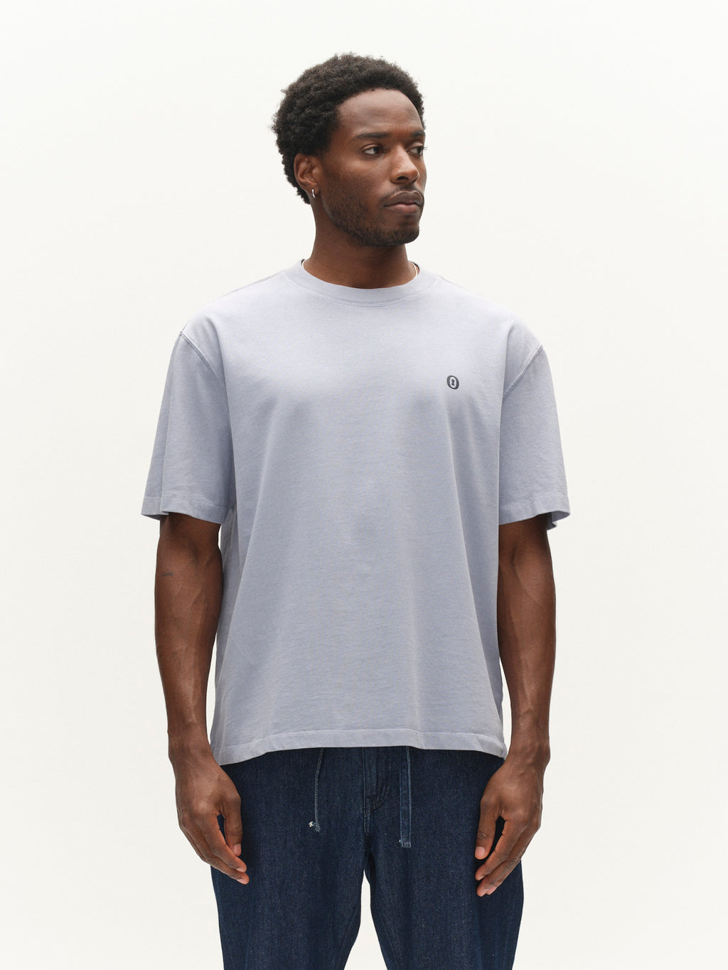 A man wearing the Gabba GABlegin Wix Boxy Hour Lobby1 SS T-shirt in dusty blue, crafted from textured cotton jersey with a small circular logo, paired with dark blue jeans, stands against a plain white background.