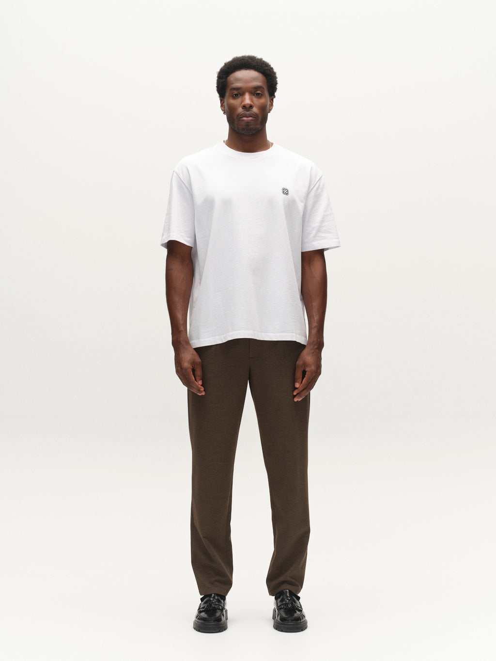 A man stands facing forward against a plain white background, wearing the Gabba GABlegin Wix Boxy Hour Lobby2 SS T-shirt in white—featuring a relaxed boxy fit and crafted from textured cotton jersey—paired with brown pants and black shoes.