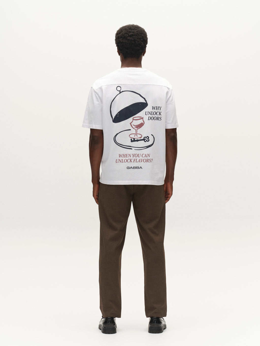 A person stands facing away, wearing the Gabba GABlegin Wix Boxy Hour Lobby2 SS T-shirt in white—a textured cotton jersey tee featuring a graphic back print of a covered dish, wine glass, and the text “WHY UNLOCK DOORS WHEN YOU CAN UNLOCK FLAVORS? GABBA.” The relaxed boxy fit is styled with brown pants and black shoes.