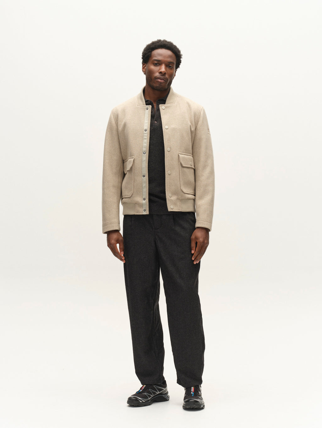 A man stands against a plain white background wearing the Gabba GABmatheo Plain Bomber Jacket in Sand, styled with a black shirt, loose black pants, and black sneakers. His calm expression and relaxed posture underscore the effortless appeal of this casual outerwear piece.