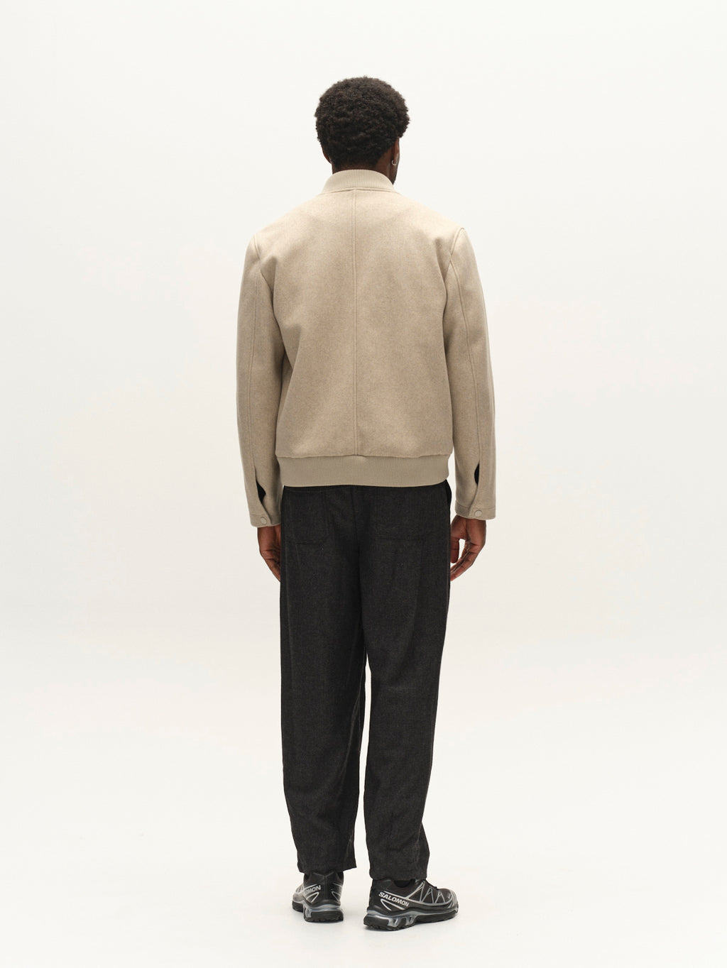 A person stands facing away against a plain light background, wearing the Gabba GABmatheo Plain Bomber Jacket in Sand, paired with dark loose pants and black sneakers.
