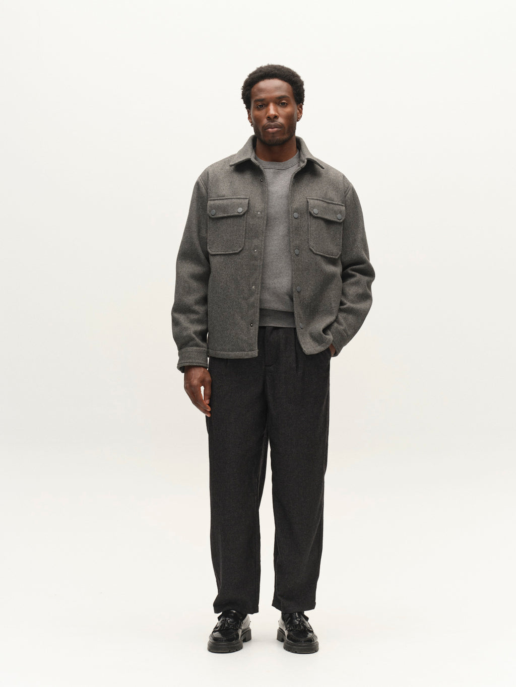 Against a plain light background, a man showcases the Gabba GABtheo City Jacket in dark grey—an overshirt-style piece featuring front pockets and wool-inspired fabric. He completes his look with a gray sweater, dark trousers, and black shoes for versatile, effortless layering.