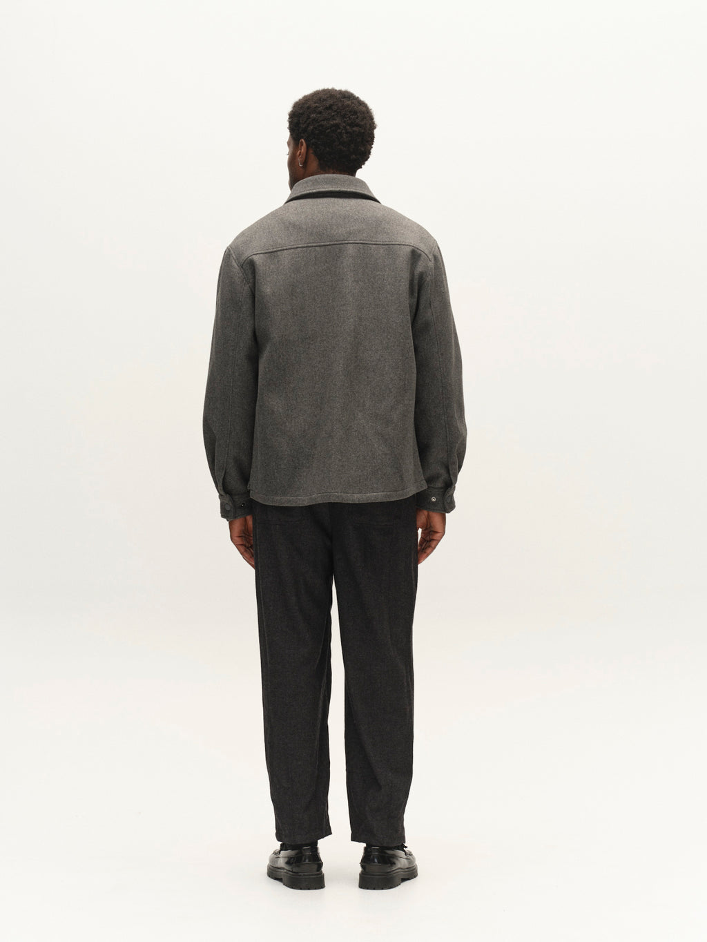 A person stands facing away from the camera, wearing the Gabba GABtheo City Jacket in Dark Grey—crafted from wool-inspired fabric—paired with dark pants and black shoes, set against a plain light background, creating an ideal outfit for everyday layering.
