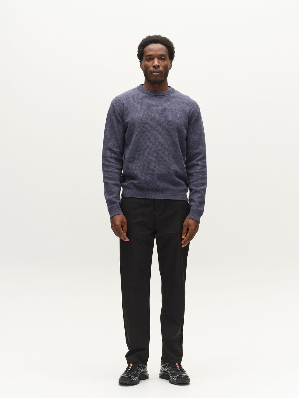 A man stands facing forward against a plain white background, wearing a light grey Gabba GABlamix Coro O-Neck LS Knit sweater crafted from a soft cotton-blend knit, paired with black pants and black athletic shoes.