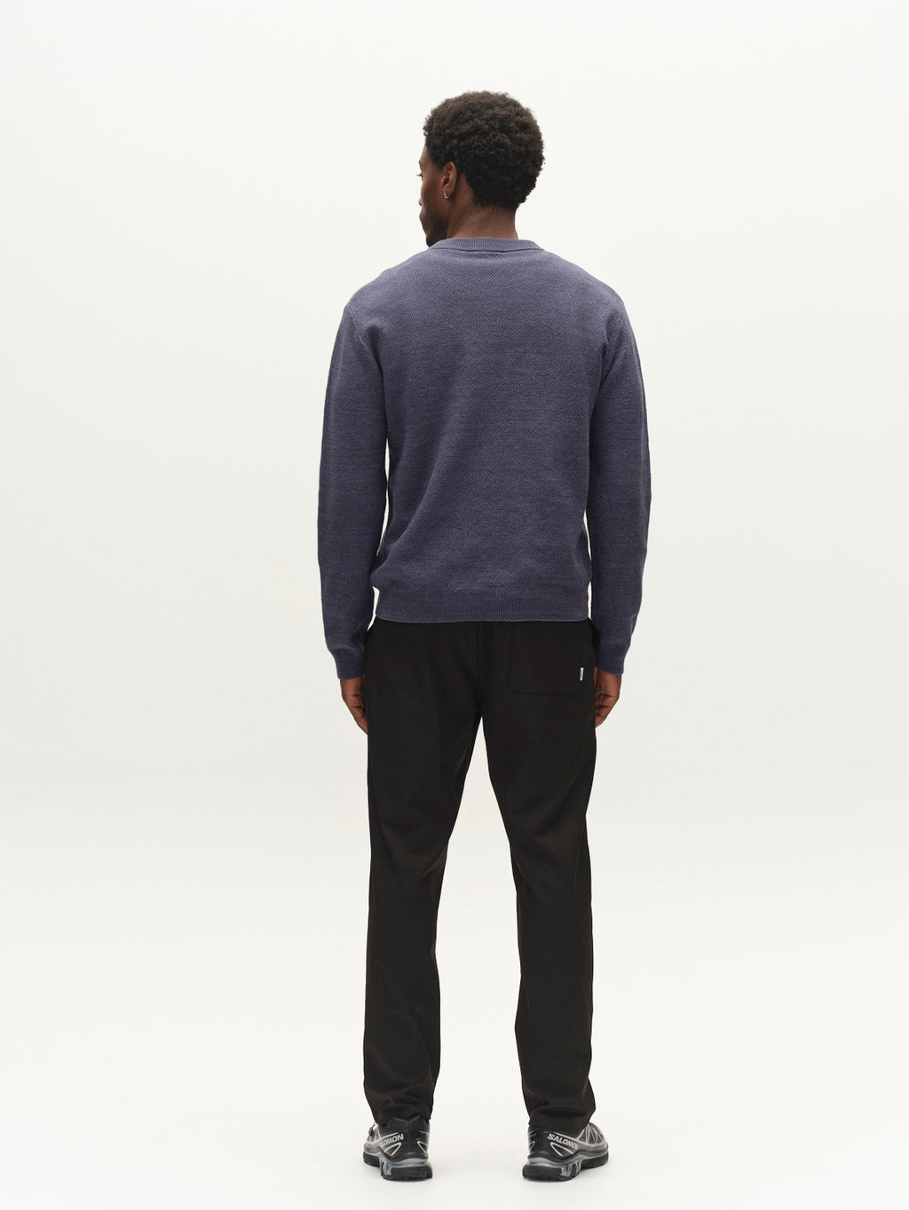 A person with short curly hair, wearing the Gabba GABlamix Coro O-Neck LS Knit in light grey—a long-sleeve crew neck sweater made from a soft cotton-blend knit—paired with black pants and gray sneakers, stands facing away from the camera against a plain white background.
