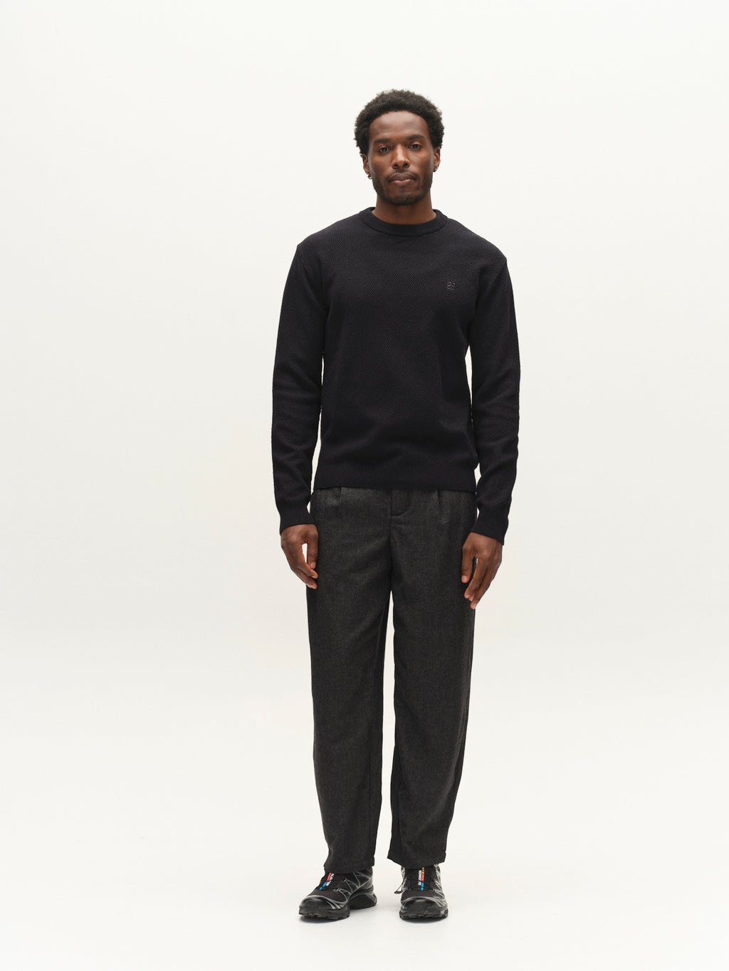 A man stands facing the camera against a plain white background, wearing the Gabba GABlamix Coro O-Neck LS Knit in Blue Black, paired with dark gray pants and black sneakers.
