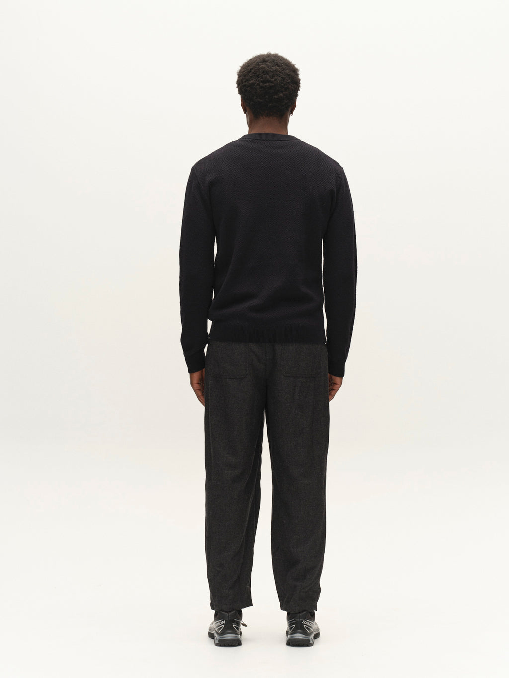 A person with short curly hair, wearing the Gabba GABlamix Coro O-Neck LS Knit sweater in blue black, paired with dark loose pants and black shoes, stands facing away against a plain white background.