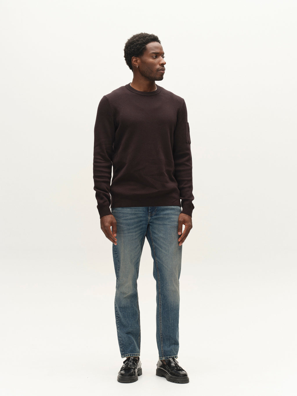 A man stands against a plain white background, wearing a dark long-sleeve sweater, Gabba GABmarc Silvi DS0007 Jeans in Mid Blue Denim crafted from comfort stretch fabric, and black shoes. He is looking to his left with his arms relaxed by his sides.