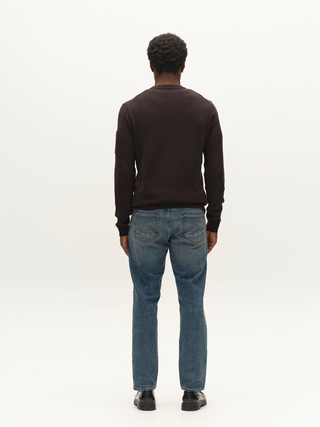 A man stands facing away from the camera against a plain white background, wearing a dark long-sleeve shirt, black shoes, and Gabba GABmarc Silvi DS0007 Jeans in mid blue denim crafted from comfort stretch fabric.