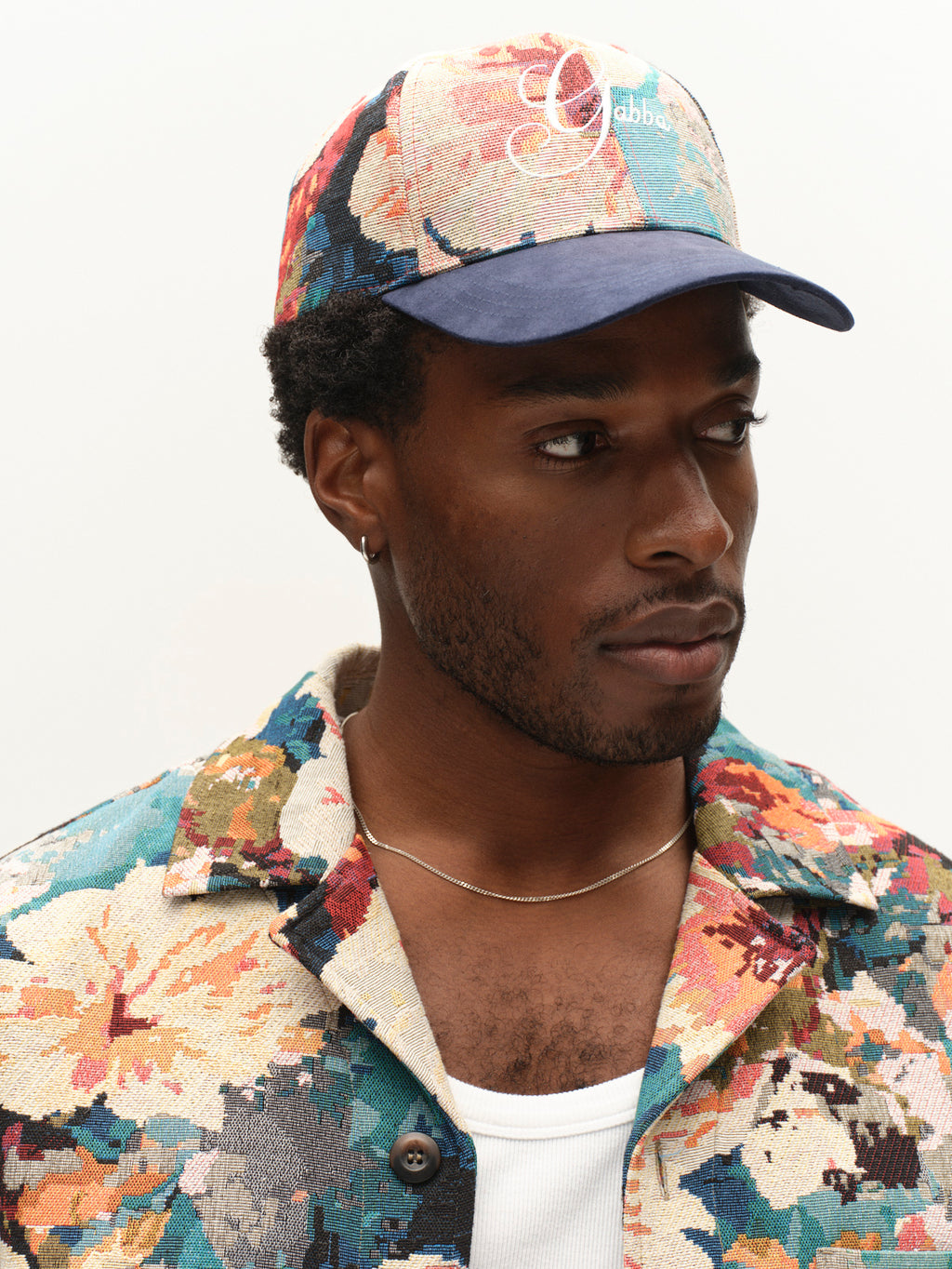A man with short hair and a trimmed beard gazes to the side, dressed in a white shirt and thin chain necklace. He wears a colorful floral-patterned jacket featuring an embroidered Gabba logo, paired with the Gabba GABrichi Fiore Cap in Multi Pattern.