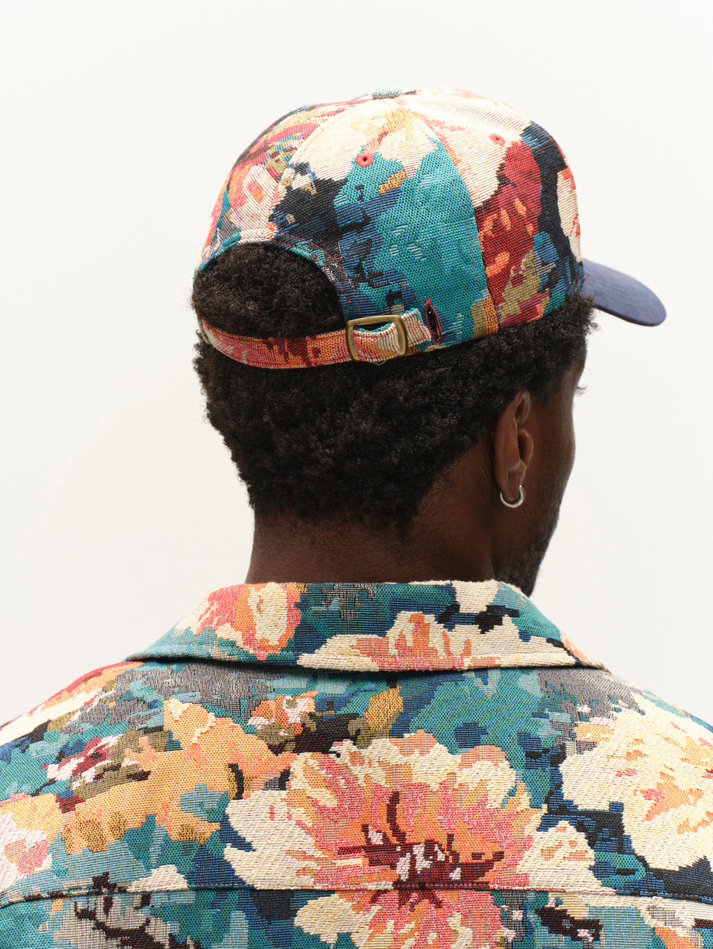 A person facing away, wearing a vibrant, floral-patterned jacket and the Gabba GABrichi Fiore Cap – Multi Pattern, featuring an adjustable back strap. Only the back of their head and shoulders are visible, with a short haircut and a hoop earring. The cap subtly displays the embroidered Gabba logo.