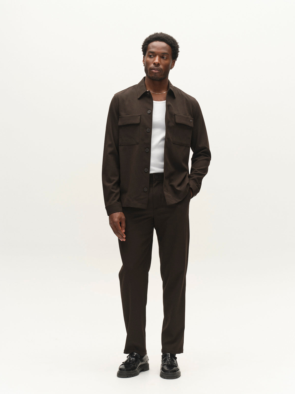 A man stands against a plain white background wearing the Gabba GABtogger Push Phin LS Overshirt GRS in Brown Slate, styled with matching pants, a white T-shirt, and black shoes. He has one hand in his pocket and gazes slightly to his left.