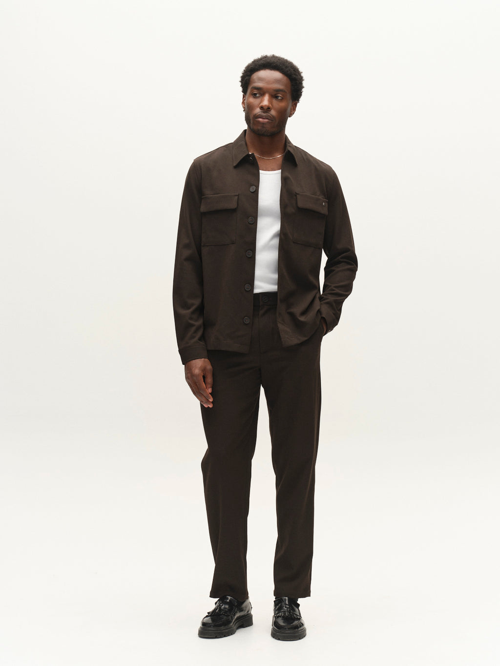 A man stands against a plain white background wearing the Gabba GABtogger Push Phin LS Overshirt GRS in Brown Slate, styled with matching pants, a white T-shirt, and black shoes. He has one hand in his pocket and gazes slightly to his left.