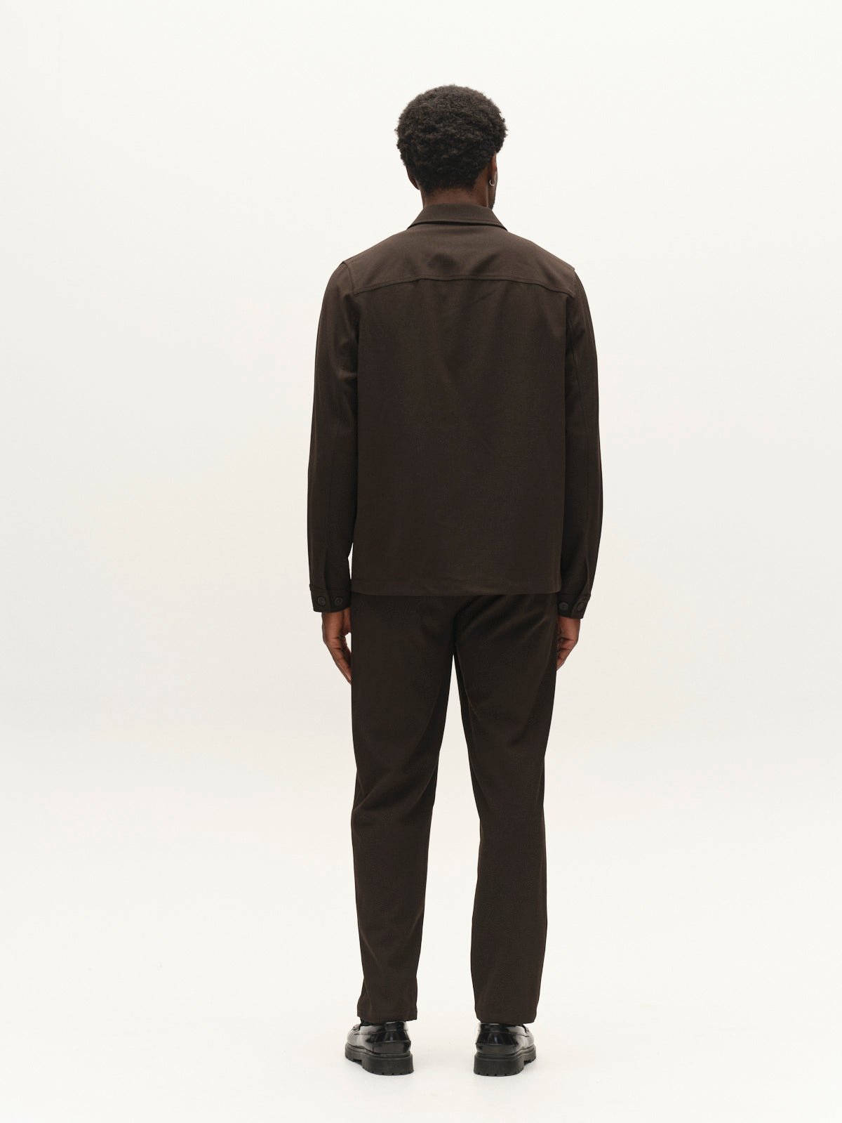 A man with short curly hair, dressed in the Gabba GABtogger Push Phin LS Overshirt GRS in Brown Slate and matching trousers, stands facing away from the camera against a plain white background. Crafted from a premium wool-viscose blend in a regular fit, this ensemble is a versatile layering essential for any modern wardrobe.