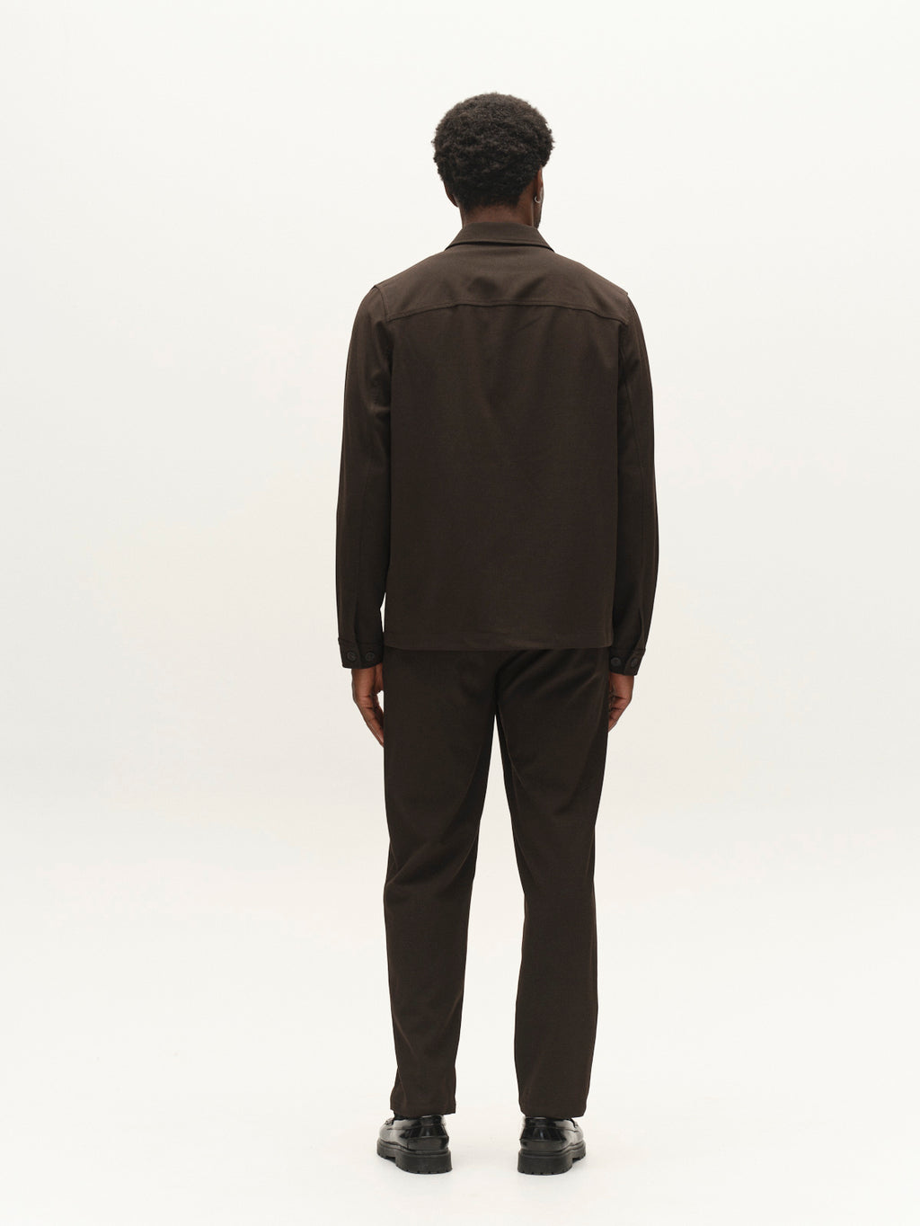 A man with short curly hair, dressed in the Gabba GABtogger Push Phin LS Overshirt GRS in Brown Slate and matching trousers, stands facing away from the camera against a plain white background. Crafted from a premium wool-viscose blend in a regular fit, this ensemble is a versatile layering essential for any modern wardrobe.