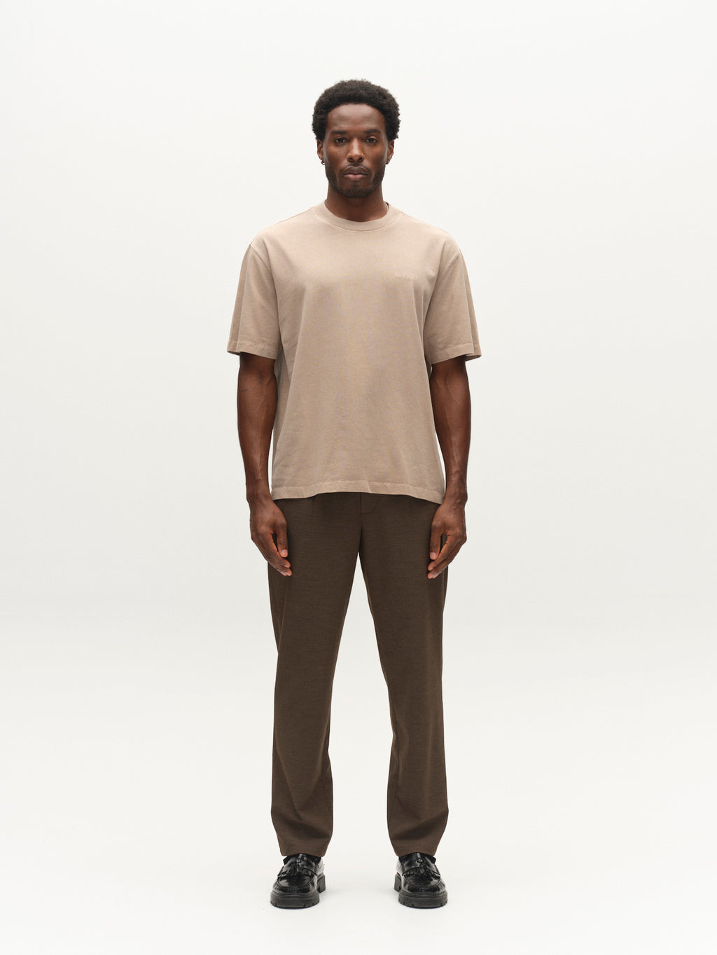 A man stands facing forward, wearing the Gabba GABlegin Wix Boxy Hour SS T-shirt in white—a relaxed, boxy-fit cotton jersey tee featuring an embroidered Gabba logo—as well as brown pants and black shoes, set against a plain white background.