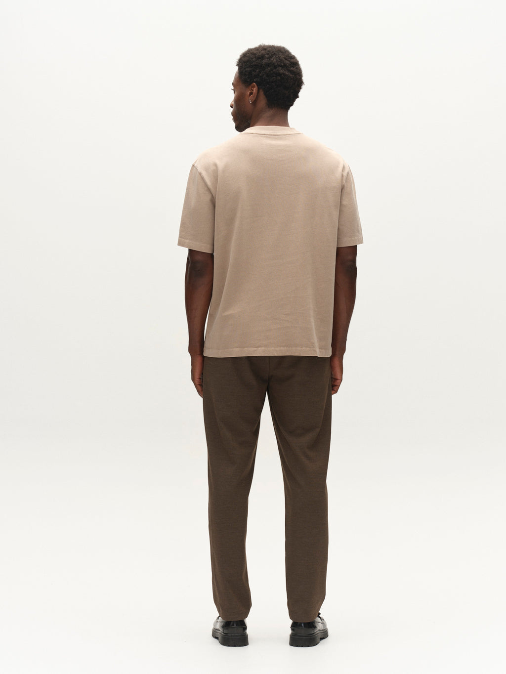 A man stands facing away, wearing the Gabba GABlegin Wix Boxy Hour SS T-shirt in white, which features a relaxed boxy fit and an embroidered Gabba logo. He pairs it with brown pants and black shoes, while the background is plain and light-colored.