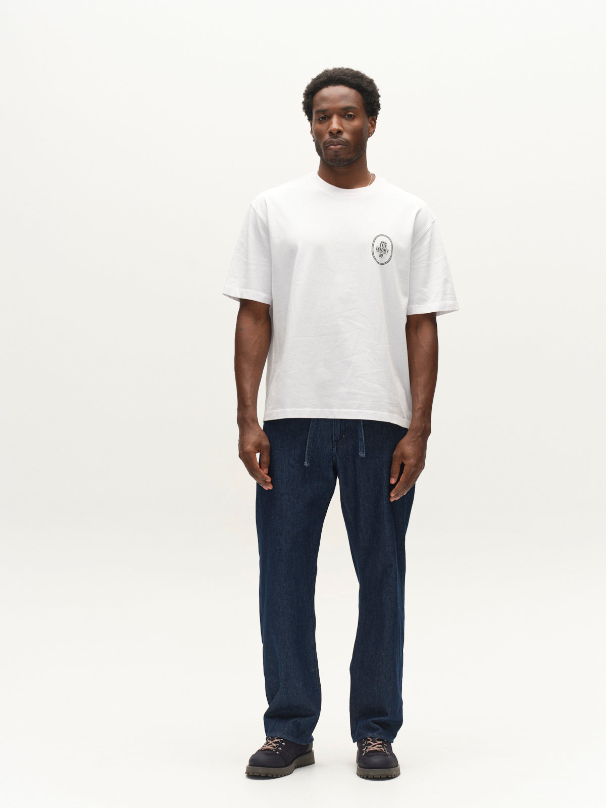 A man stands against a plain white background, wearing the Gabba GABlegin Boxy Fiore SS T-Shirt in white—a relaxed, boxy-fit short-sleeve tee crafted from textured cotton jersey featuring a small logo on the chest. He pairs it with dark blue pants and black shoes, facing forward with a neutral expression and embodying an effortlessly stylish contemporary wardrobe staple.