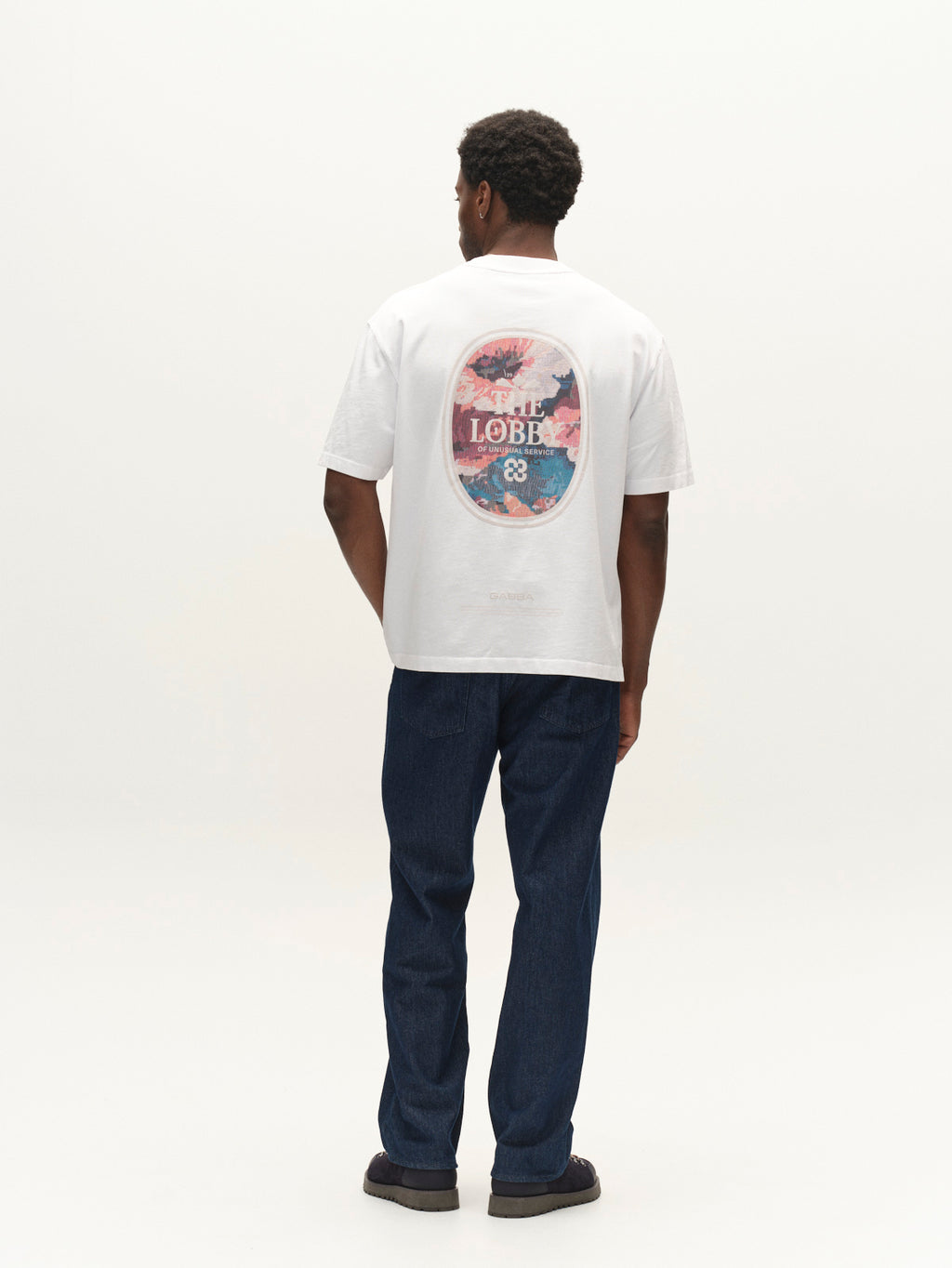 A man stands facing away, wearing the Gabba GABlegin Boxy Fiore SS T-Shirt in white—a relaxed, boxy-fit cotton jersey tee featuring a colorful circular graphic and text on the back—paired with blue jeans and black shoes against a plain white background. This contemporary wardrobe staple offers both comfort and style.