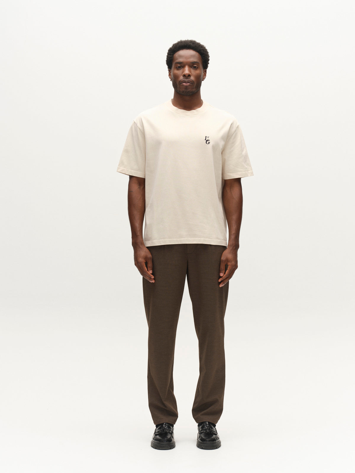 A man stands facing forward against a plain white background, wearing the Gabba GABlegin Boxy Glory SS T-Shirt in Birch—a beige, short-sleeve tee made from textured cotton jersey with a small logo on the left chest—paired with brown pants and black shoes.