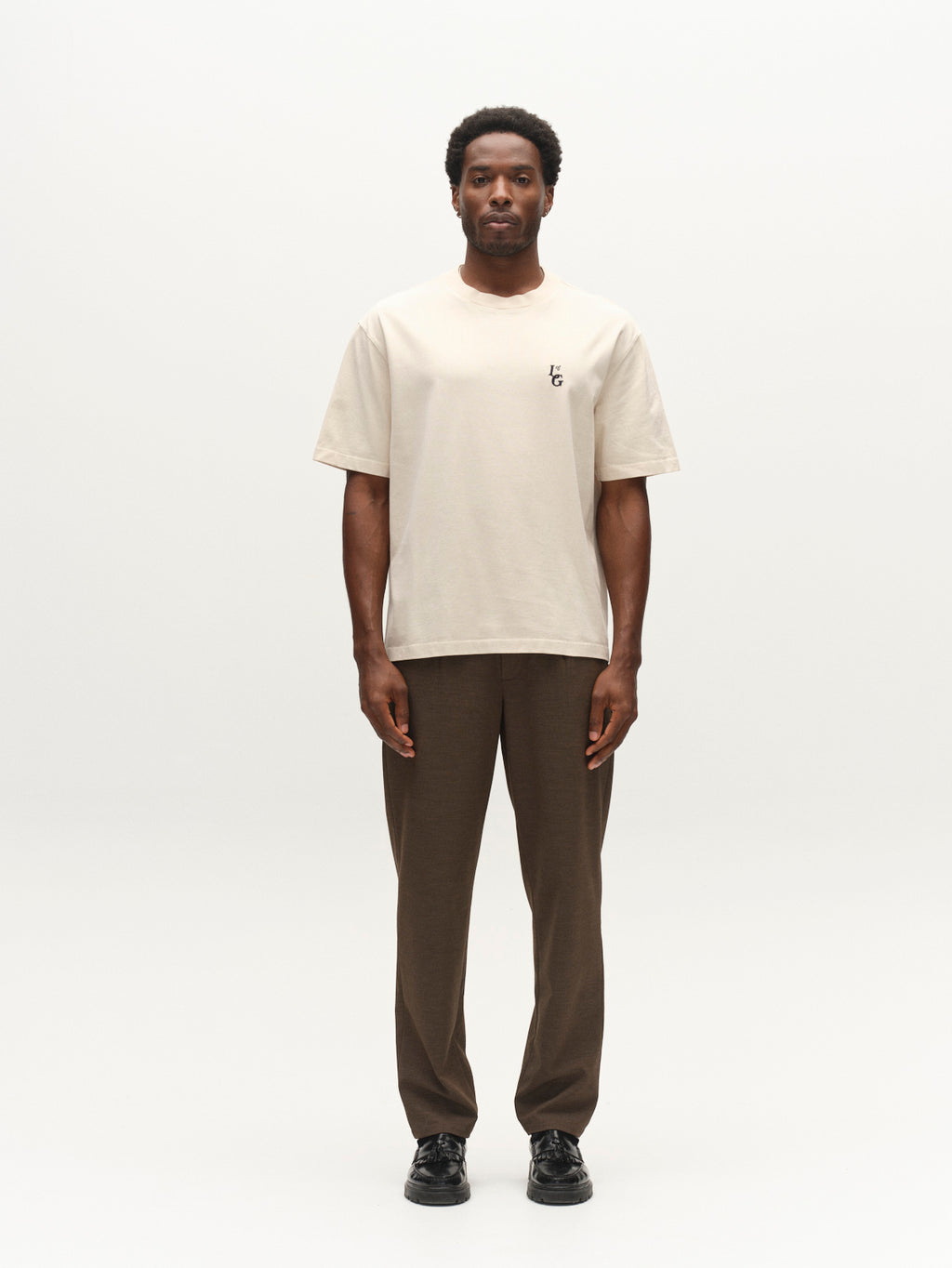 A man stands facing forward against a plain white background, wearing the Gabba GABlegin Boxy Glory SS T-Shirt in Birch—a beige, short-sleeve tee made from textured cotton jersey with a small logo on the left chest—paired with brown pants and black shoes.