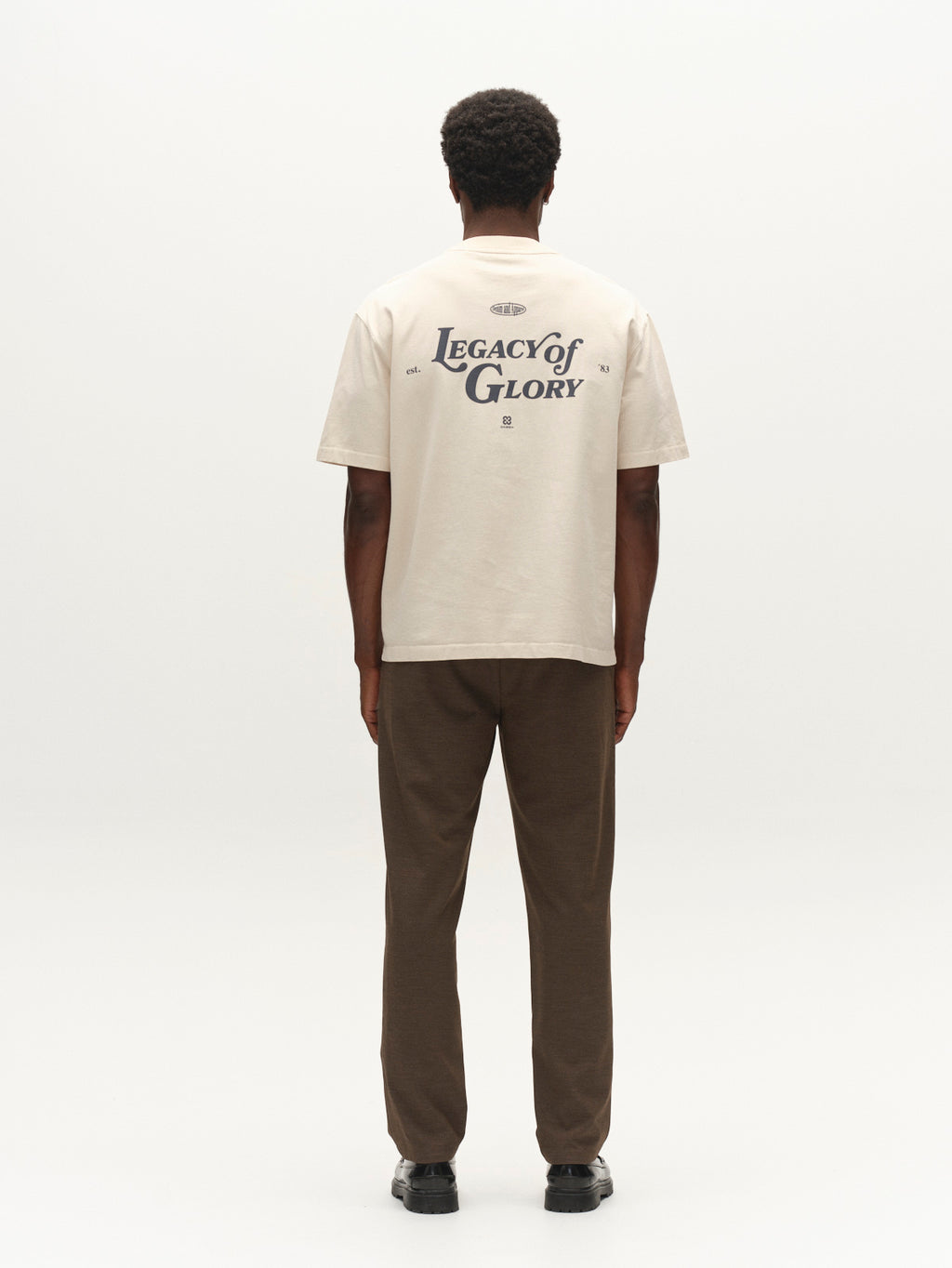 A person stands facing away, wearing the Gabba GABlegin Boxy Glory SS T-Shirt in Birch—a beige, textured cotton jersey top featuring a relaxed boxy fit and Legacy of Glory graphic back print—paired with brown pants and black shoes against a plain white background.