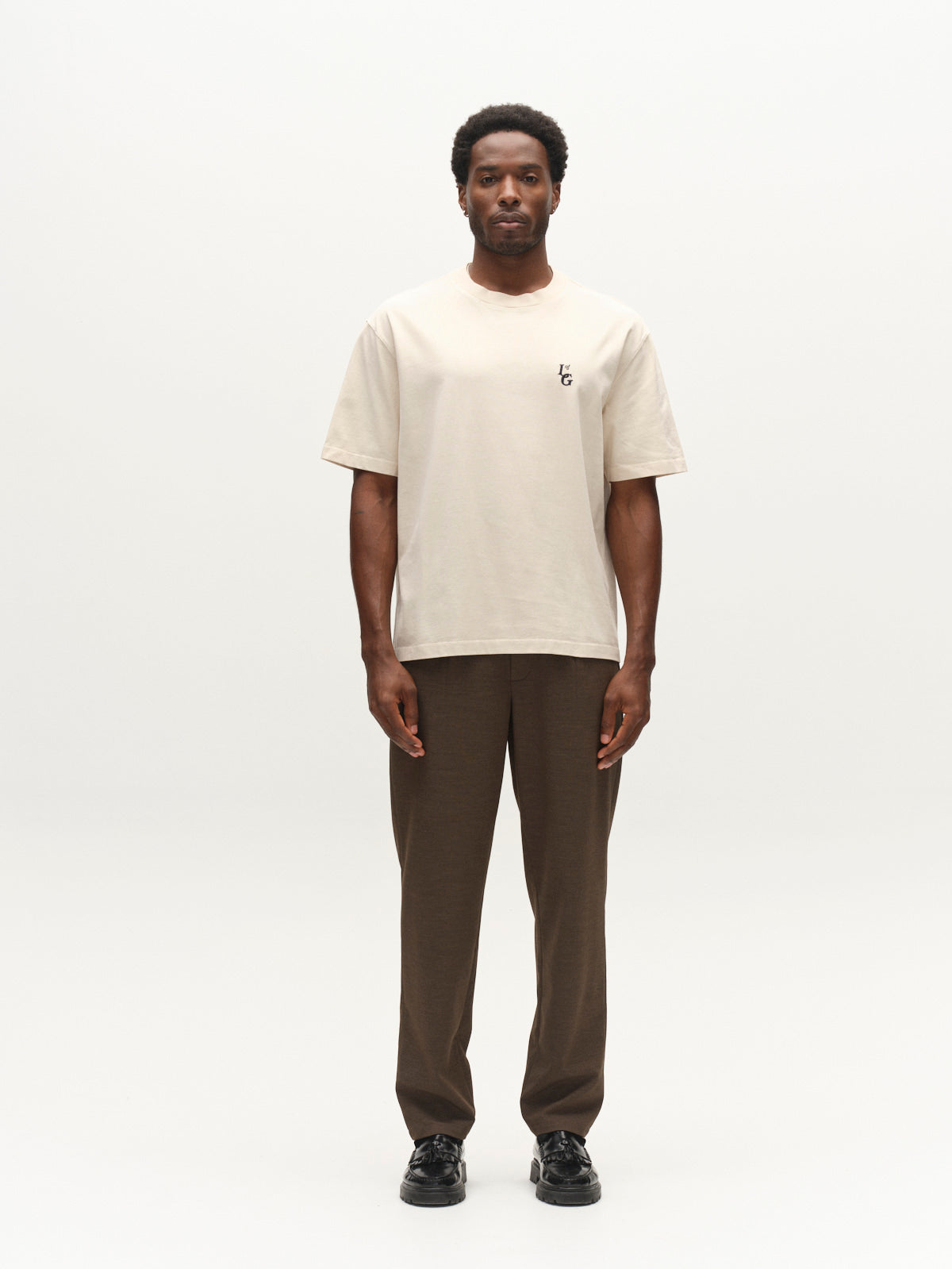A man stands facing forward against a plain white background, wearing the Gabba GABlegin Boxy Glory SS T-Shirt in Birch—a beige textured cotton jersey tee with a small black chest logo—paired with brown pants and black shoes.