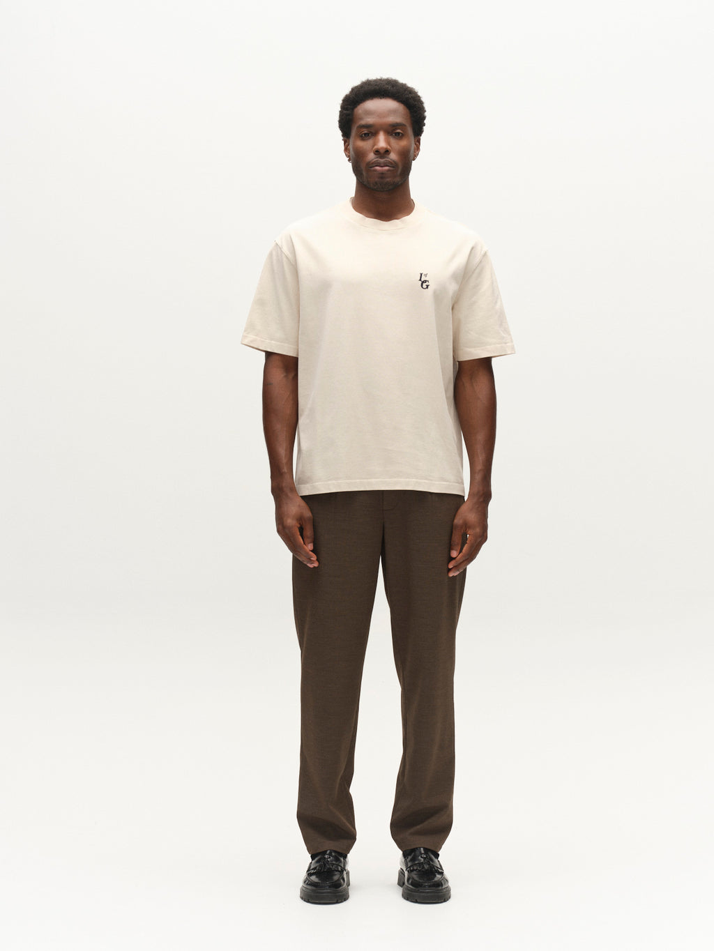 A man stands facing forward against a plain white background, wearing the Gabba GABlegin Boxy Glory SS T-Shirt in Birch—a beige textured cotton jersey tee with a small black chest logo—paired with brown pants and black shoes.