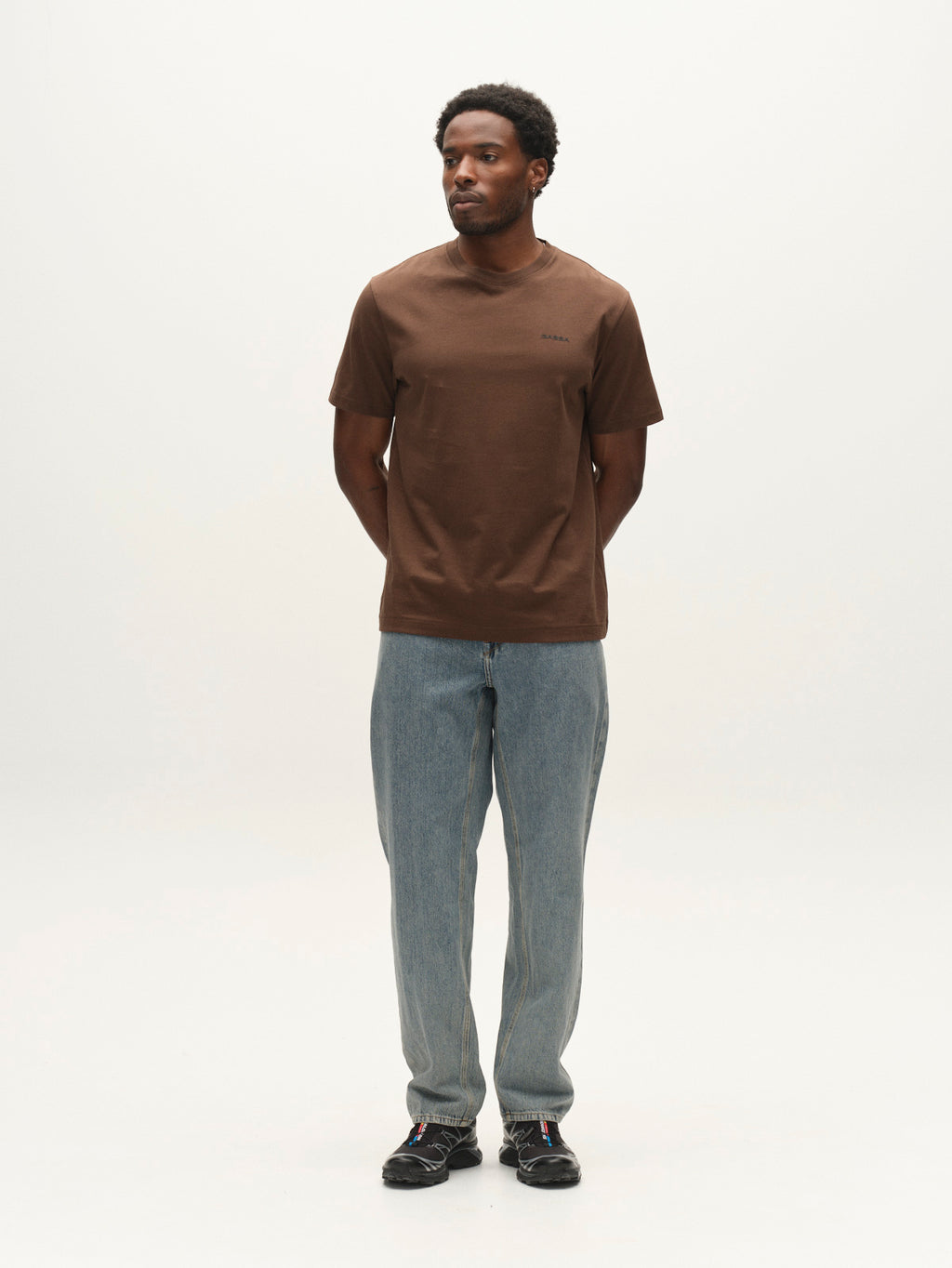 A man stands against a plain white background, wearing the Gabba GABdune Logo GOTS SS T-Shirt in Brown Slate, paired with light blue jeans and black shoes. His hands are behind his back, and he looks slightly to his left.