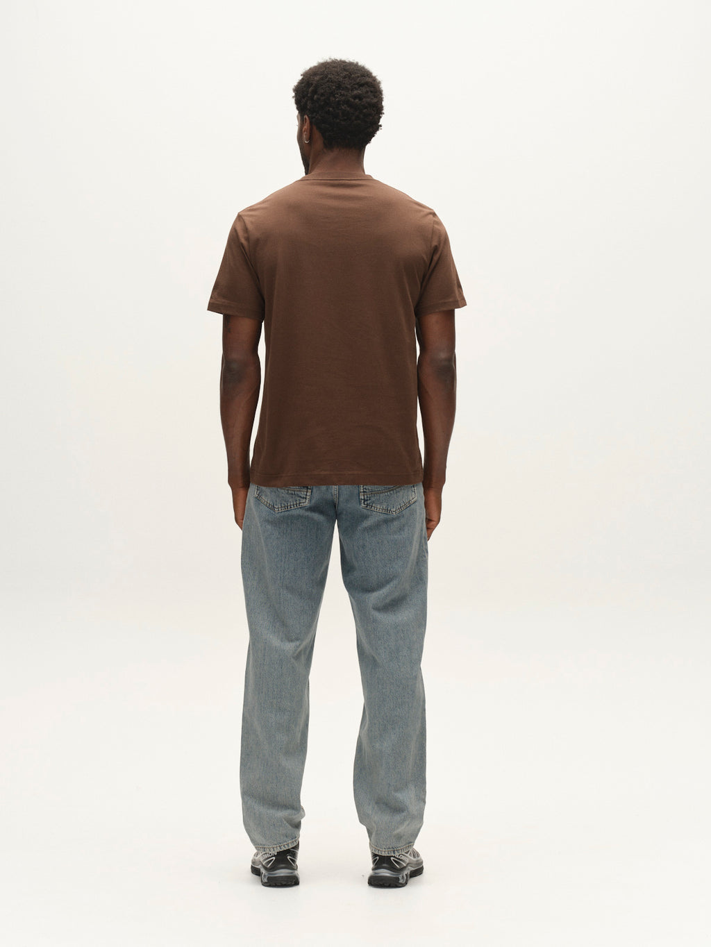 A person stands facing away from the camera, wearing the Gabba GABdune Logo GOTS SS T-Shirt in Brown Slate, light blue jeans, and black shoes, against a plain white background.