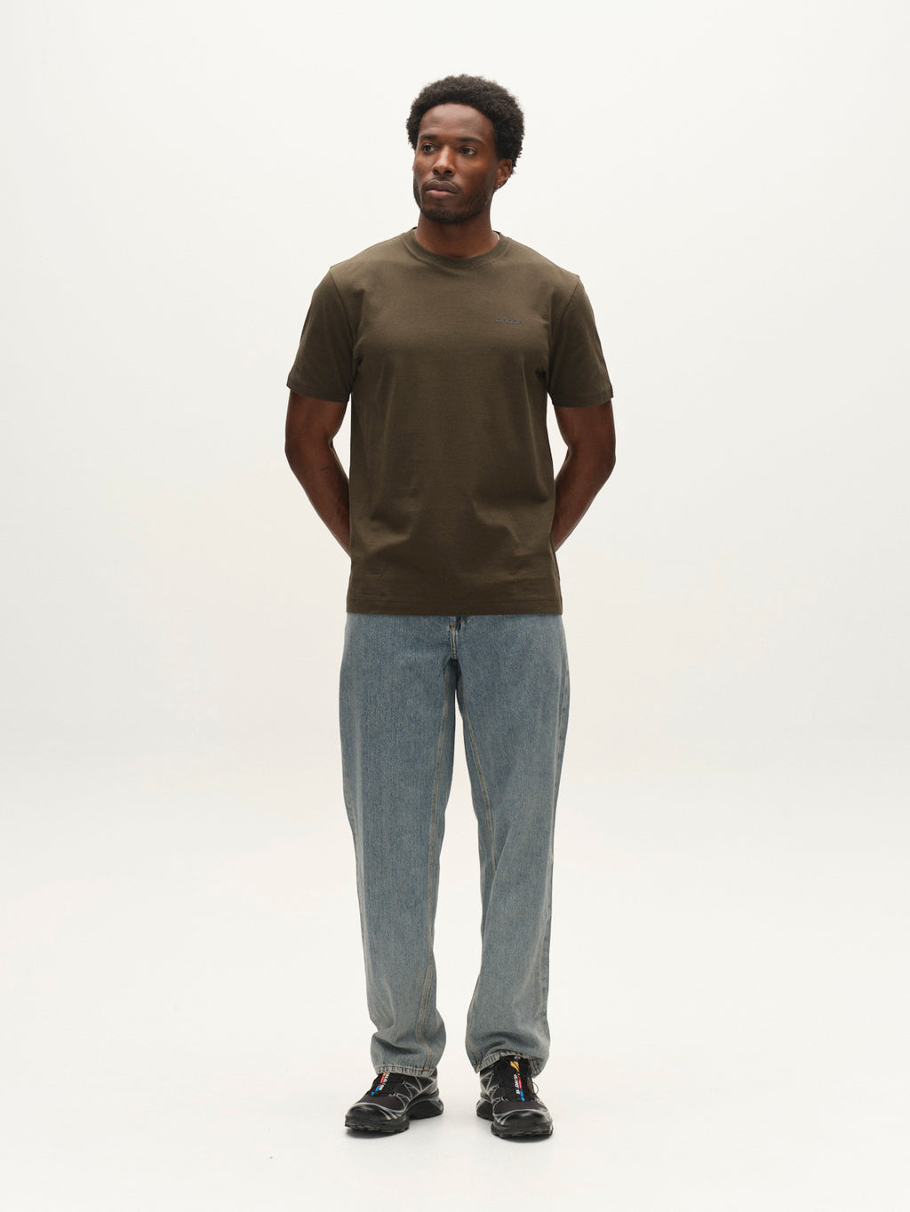 A man stands against a plain white background with his hands behind his back, wearing the Gabba GABdune Logo GOTS SS T-Shirt in Forest Night, light blue jeans, and black sneakers.