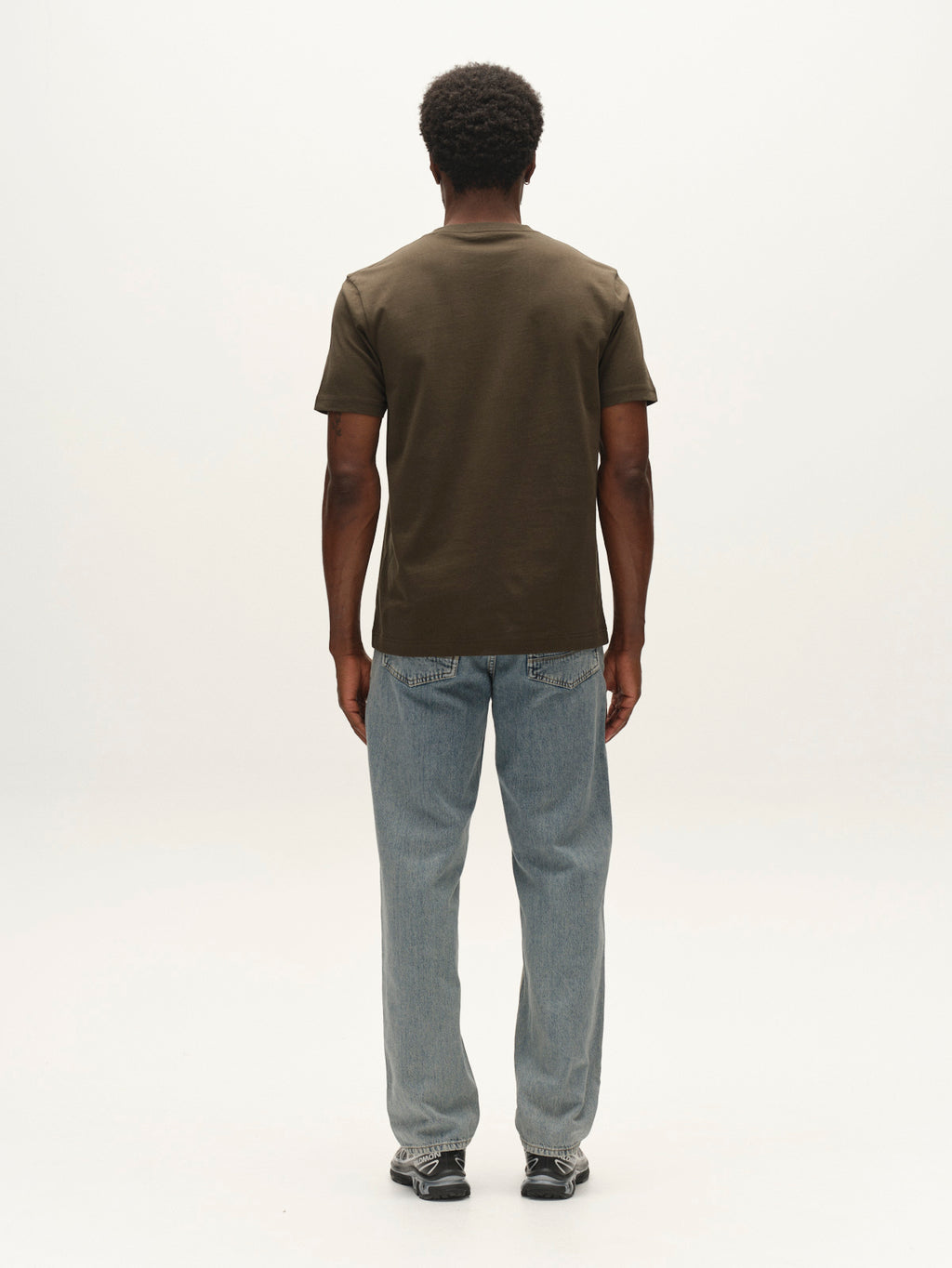 A person stands facing away from the camera, wearing a Gabba GABdune Logo GOTS SS T-Shirt in Forest Night, paired with light blue jeans and sneakers, set against a plain white background.