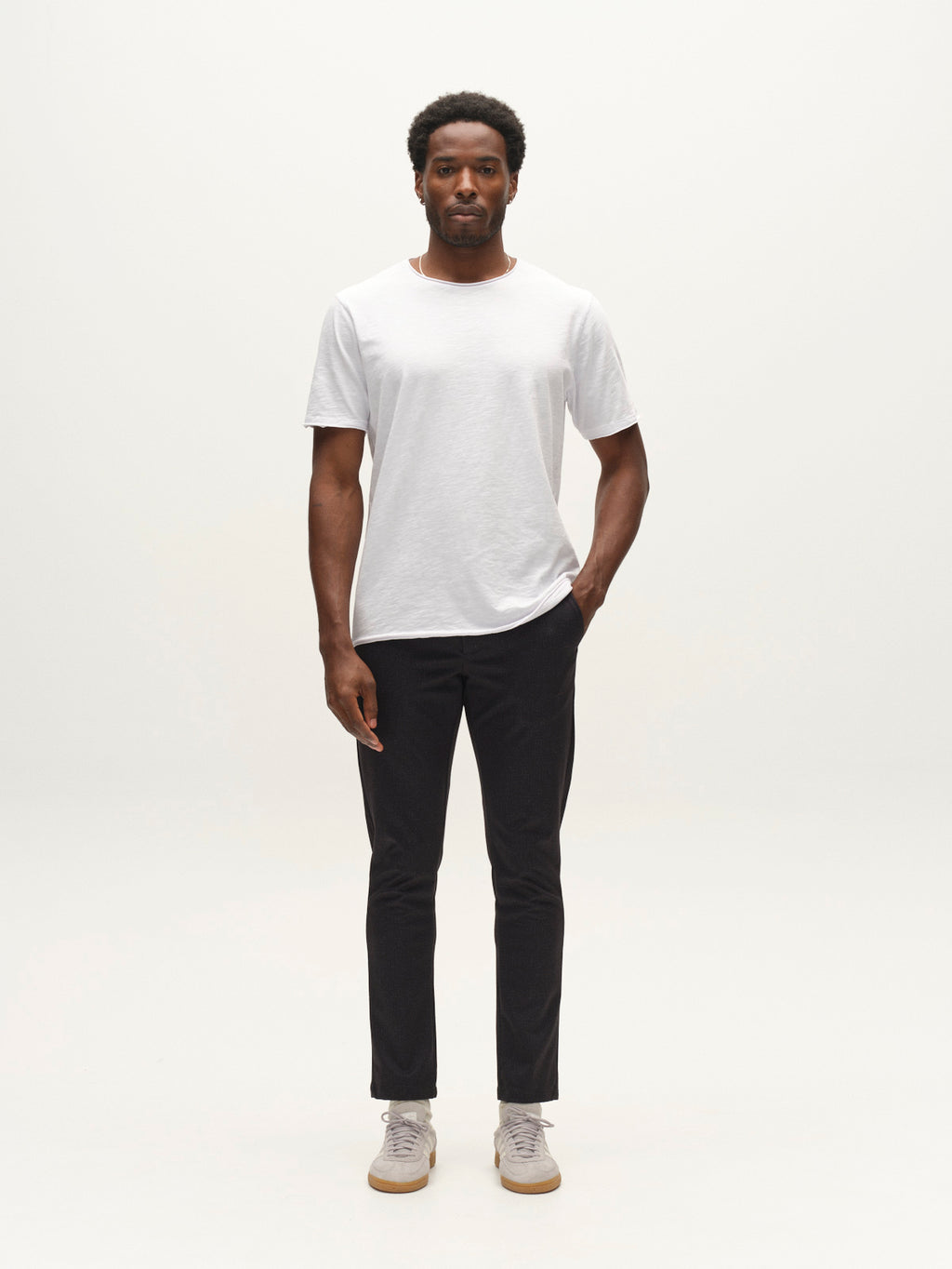 A man stands facing forward against a plain white background, wearing the GABpaul Nygo - Night Sky slim chino fit black pants from Gabba, crafted from comfort stretch fabric, paired with a white t-shirt and light-colored sneakers. One of his hands is in his pocket and he has a neutral expression.