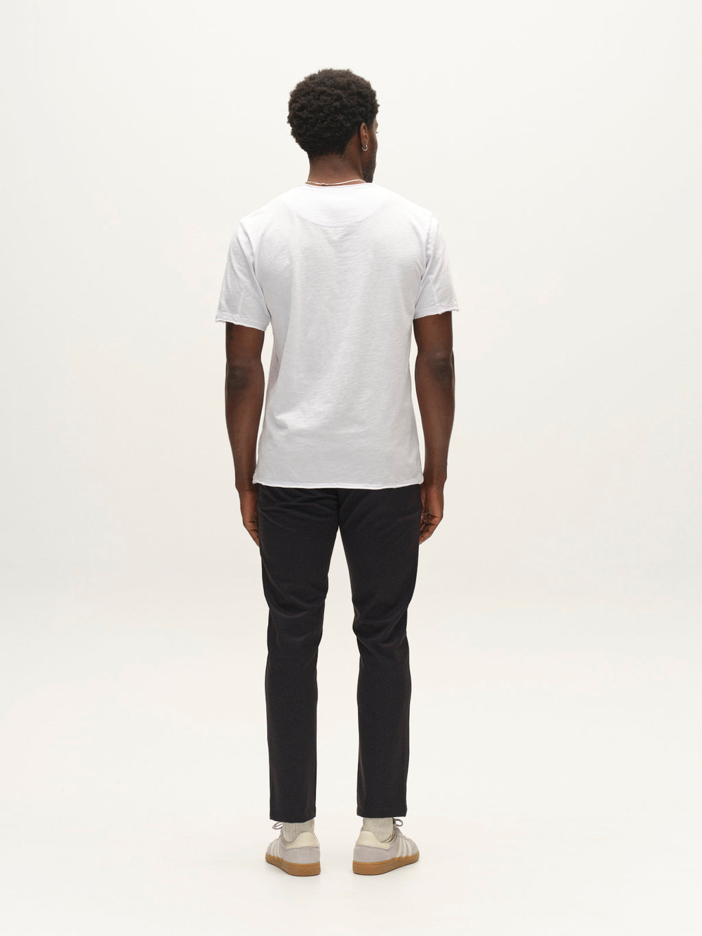 A person stands facing away from the camera, wearing a white T-shirt, light-colored shoes, and Gabba’s GABpaul Nygo - Night Sky pants crafted from comfort stretch fabric in a slim chino fit, against a plain white background.