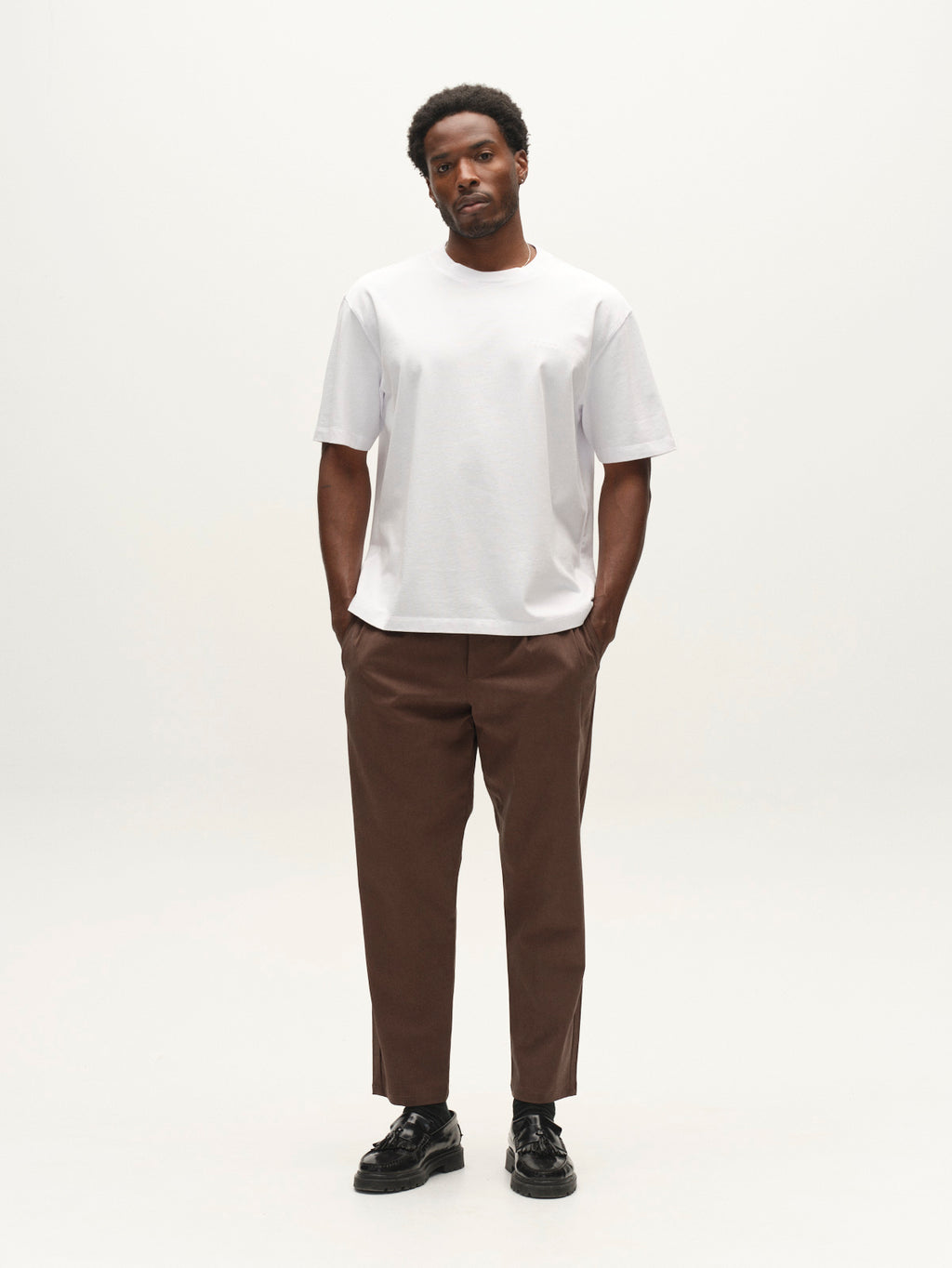 A man wears a white t-shirt, GABfirenze Bolu Cropped TENCEL ™ Pant - Brown Slate, and black loafers, standing with his hands in his pockets.