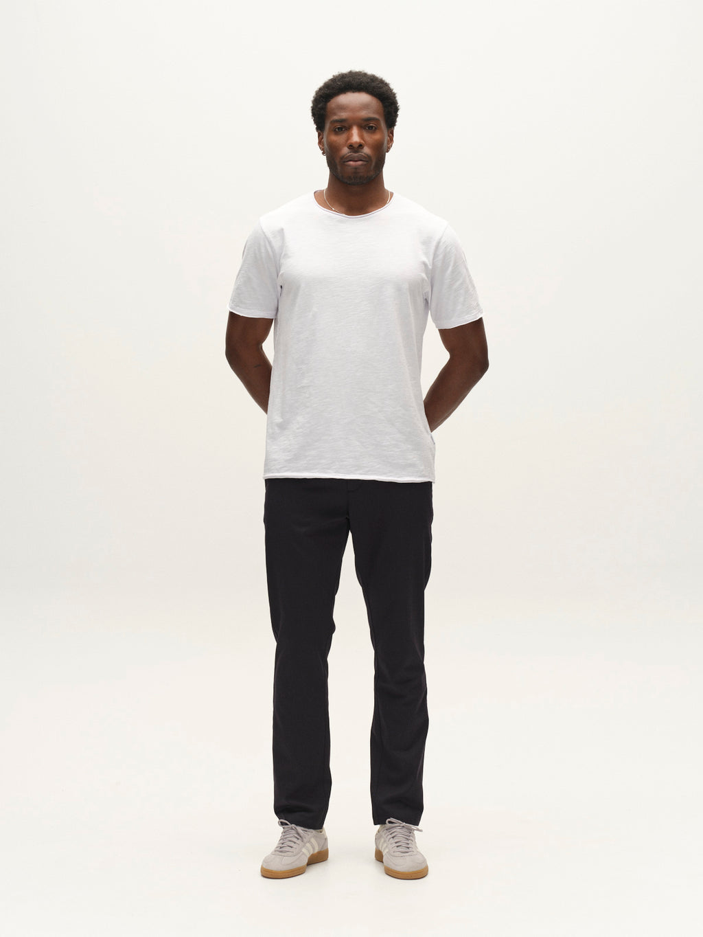 A man stands facing forward with his hands behind his back, wearing a plain white t-shirt, beige sneakers, and the Gabba GABpaul Deme Pant in Dark Navy—black slim chino fit pants crafted from micro-pattern woven fabric—against a plain white background, creating an effortless look for everyday wear.