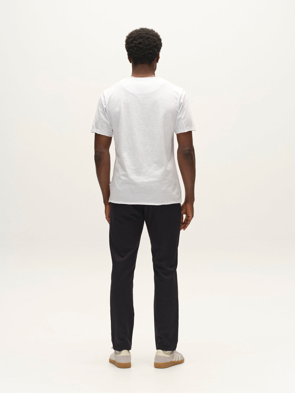 A person stands facing away, wearing a plain white T-shirt crafted from micro-pattern woven fabric, the Gabba GABpaul Deme Pant in Dark Navy with a slim chino fit, and light-colored shoes against a plain white background.