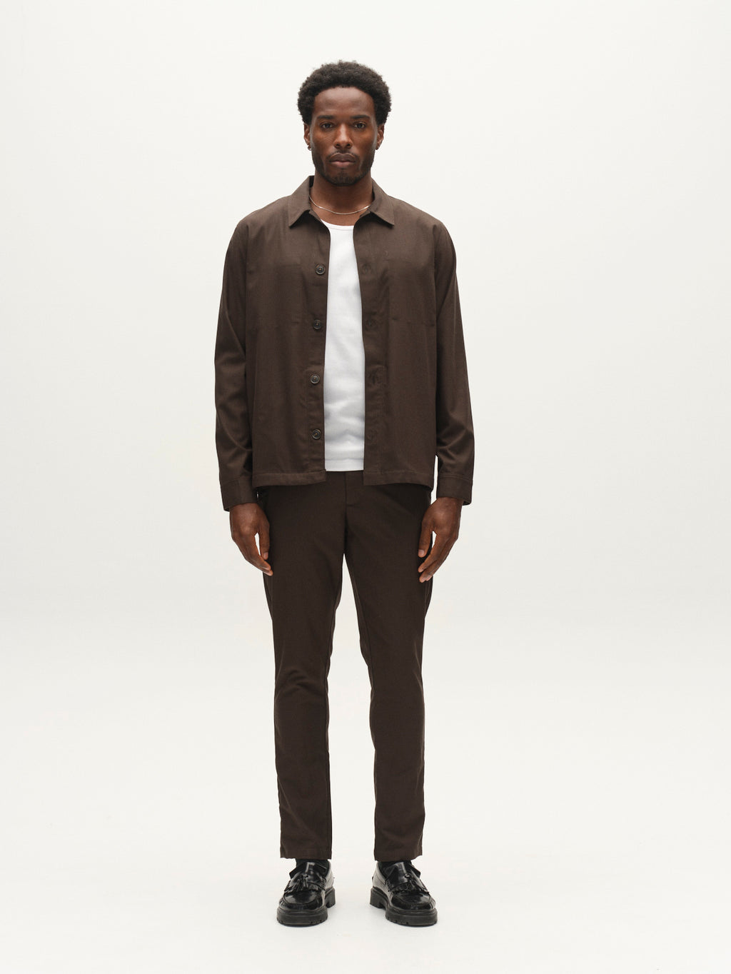 A man stands facing forward, wearing a brown button-up shirt over a white t-shirt, Gabba GABpaul Deme Pant in Forest Night with a slim chino fit, and black shoes, against a plain white background.