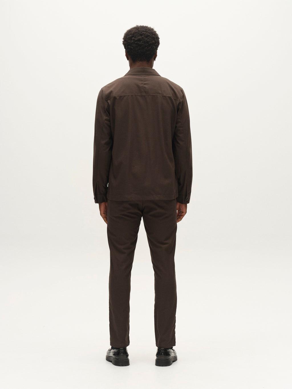 A person with short curly hair, dressed in a brown long-sleeve shirt and Gabba’s GABpaul Deme Pant in Forest Night—a slim chino fit crafted from micro-pattern woven fabric—stands facing away from the camera against a plain white background.