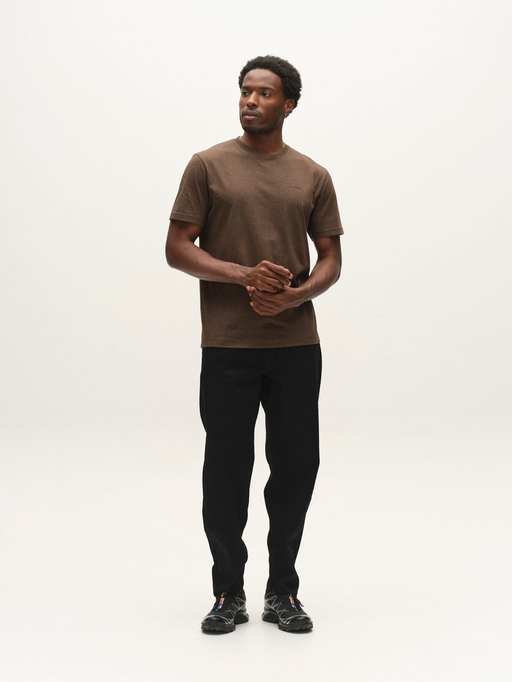 A man stands against a plain white background, wearing the Gabba GABdune Melange New SS T-Shirt in Brown Slate, paired with black pants and black shoes. He looks slightly to the side with his hands loosely clasped in front of him.